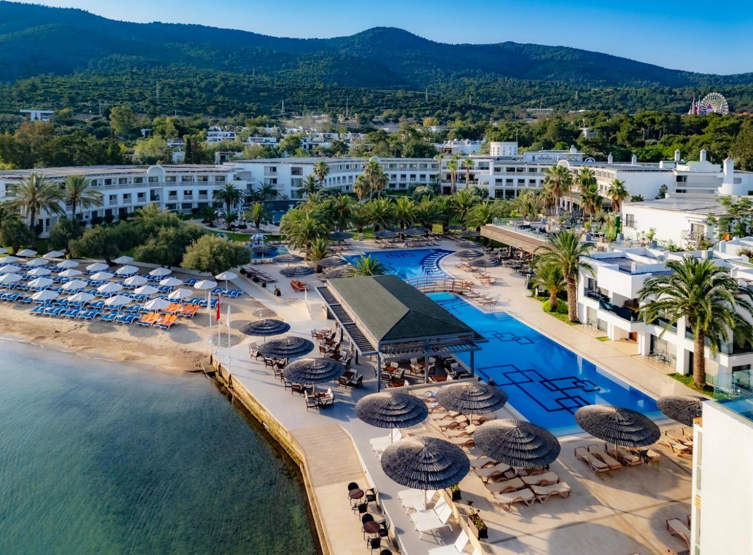 Samara Hotel Bodrum panorāmas skats ar lielu āra baseinu, privāto pludmali un kalnu fonu Turcijas piekrastē