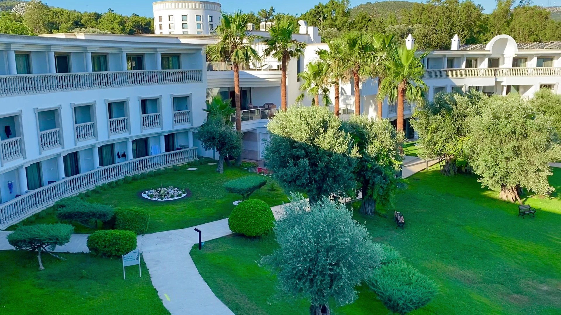 Samara Hotel Bodrum numuru korpusi ar balkoniem, ko ieskauj zaļš dārzs ar palmām un olīvkokiem