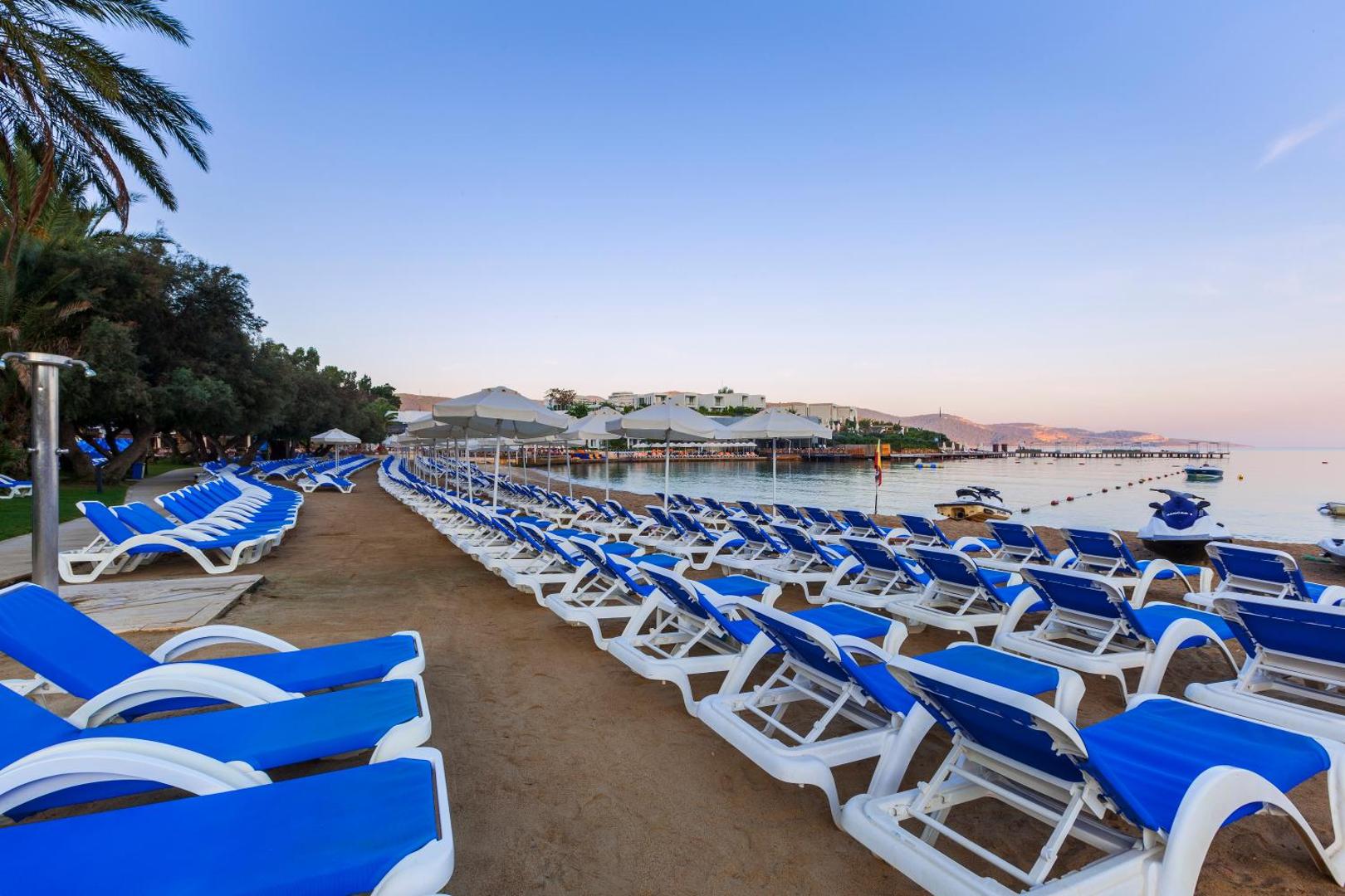Samara Hotel Bodrum plašā sauļošanās zona ar rindu zilajiem krēsliem un saulessargiem tieši pie jūras līča