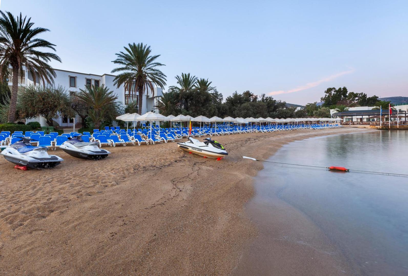 Skats uz Samara Hotel Bodrum pludmali saulrietā ar palmām, baltiem saulessargiem un ūdens motocikliem krastā