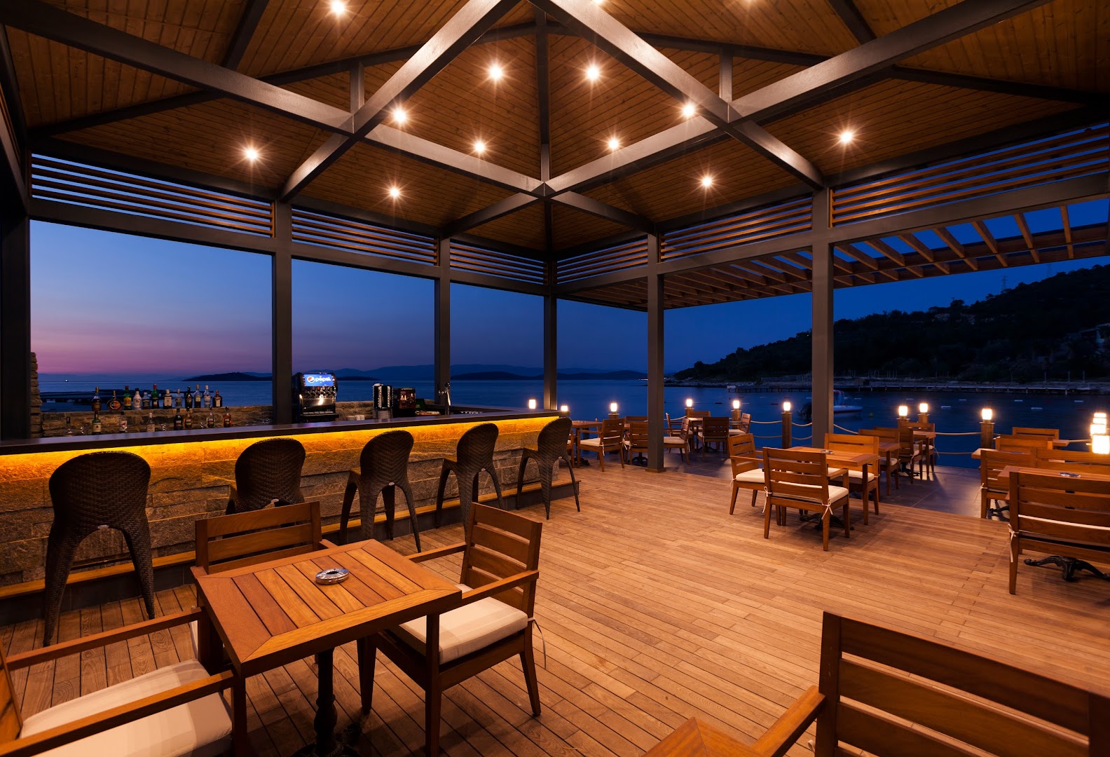 Samara Hotel Bodrum āra bārs ar koka terasi saulrieta laikā, no kura paveras skats uz Egejas jūru un piekrastes kalniem.