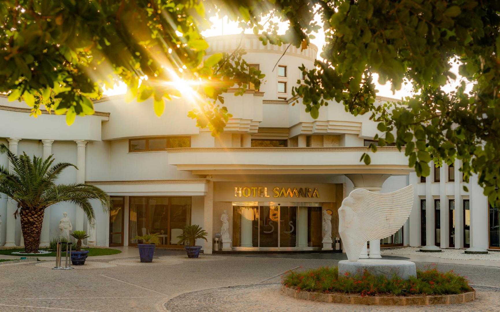 Samara Hotel Bodrum galvenā ieeja saulrieta gaismā ar baltām skulptūrām un palmām pie viesnīcas fasādes