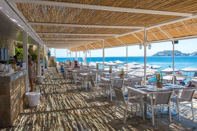 Peninsula Resort & Spa āra terases restorāns zem niedrīšu jumta ar skatu uz Vidusjūru un piekrastes klintīm