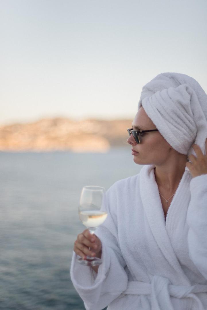 Viesis baltā peldmētelī ar vīna glāzi Peninsula Resort & Spa terasē uz saulrieta un jūras fona