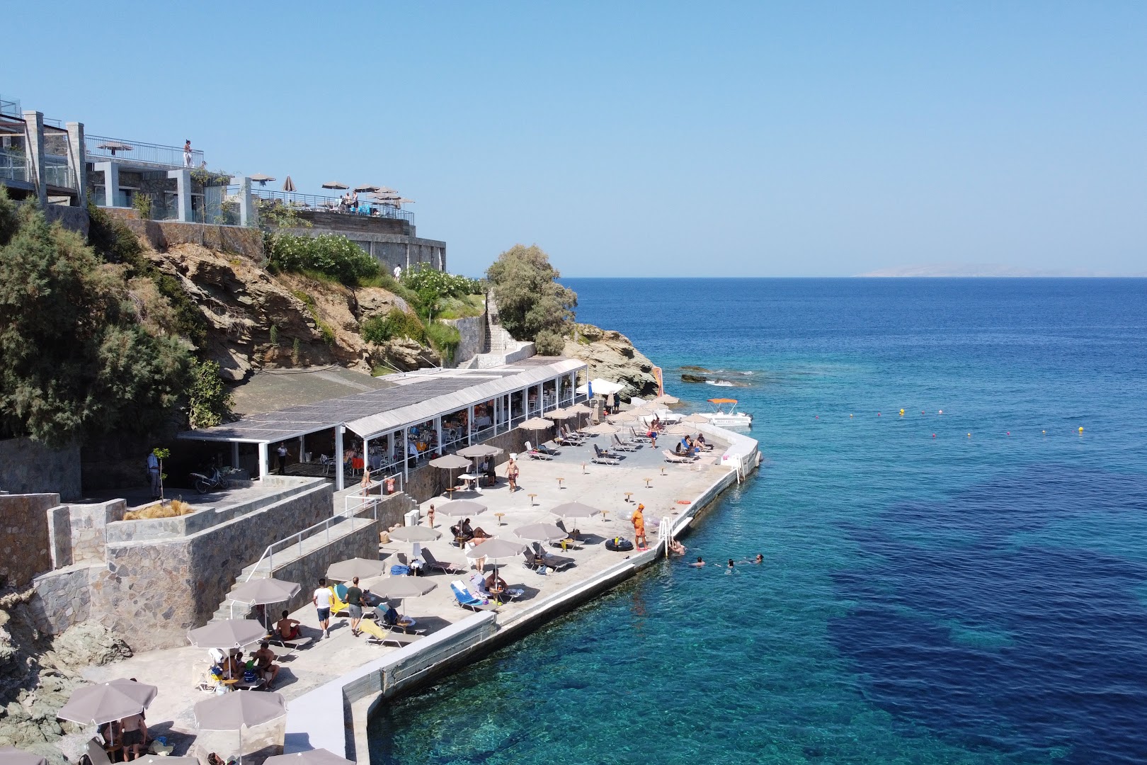 Peninsula Resort & Spa pludmales zona uz klints platformas Krētā ar saulessargiem, gultām un tiešu piekļuvi dzidram jūras ūdenim