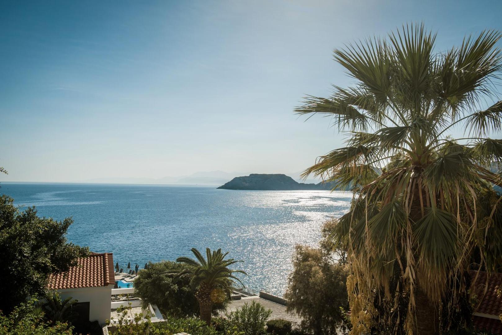 Peninsula Resort & Spa teritorija ar palmām un skatu uz jūru, saules apspīdētiem kalniem un viesnīcas ēku jumtiem.