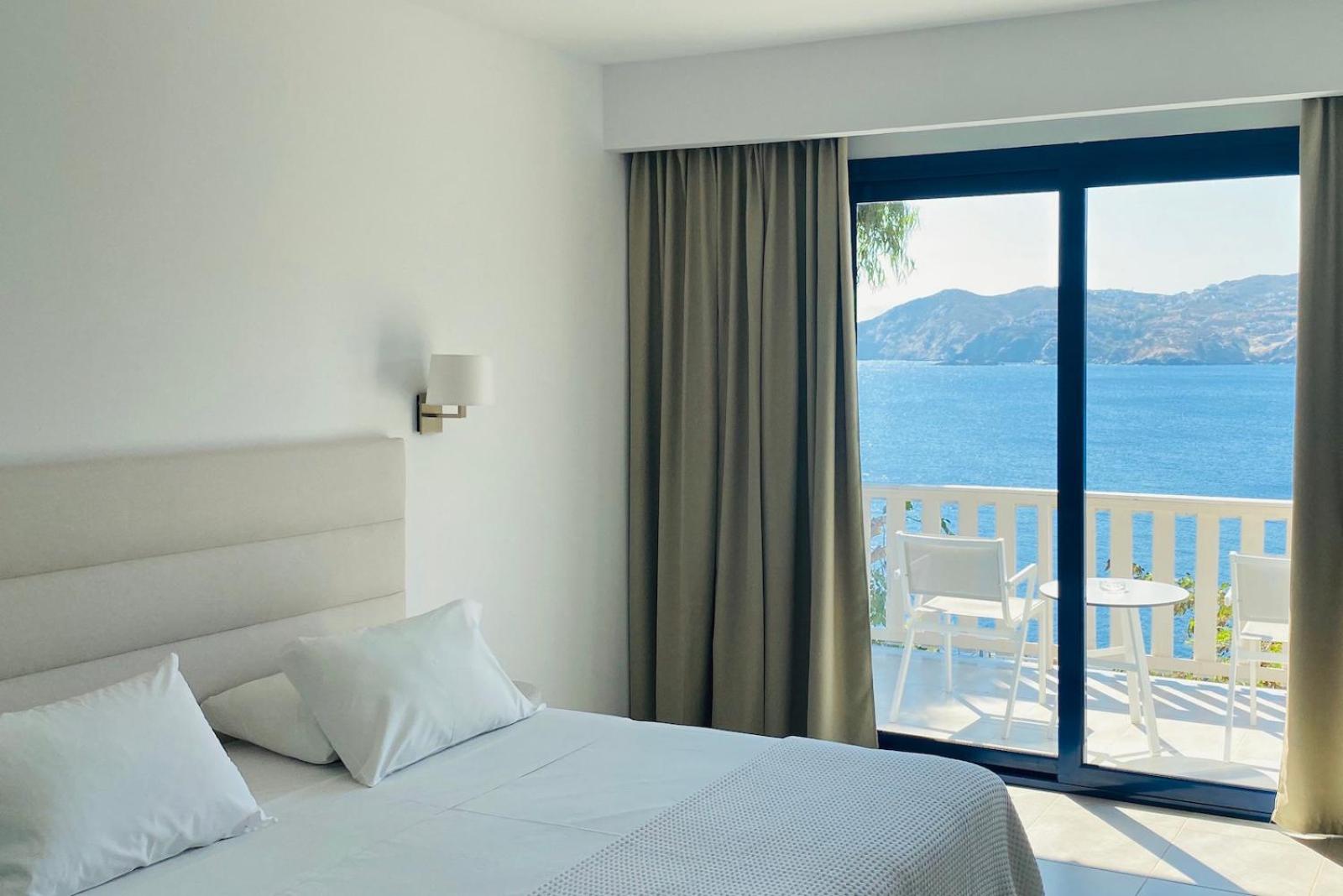 Peninsula Resort & Spa numura interjers ar skatu uz kalniem un jūru Krētā — balta gulta un balkons ar mēbelēm