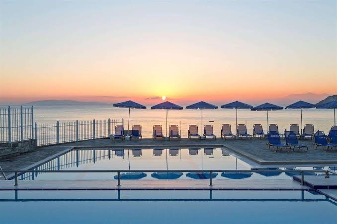 Saulriets pie Peninsula Resort & Spa baseina — rinda ar sauļošanās krēsliem un saulessargiem pretī jūras horizontam vakarā
