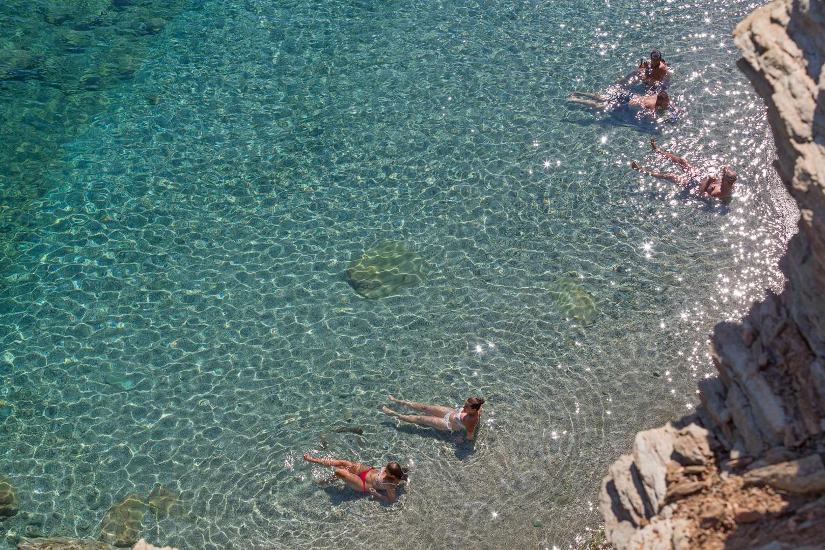 Atpūtnieki peldas un relaksējas Peninsula Resort & Spa kristāldzidrajā piekrastes ūdenī pie klintīm saulainā pēcpusdienā.