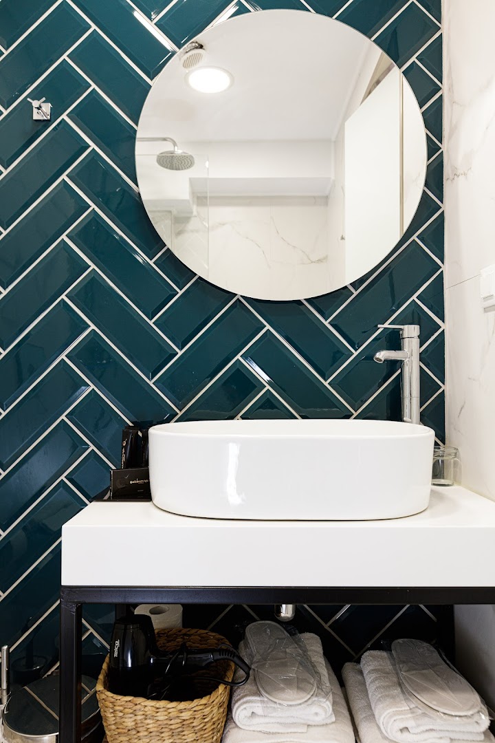 Moderna Joan Palace numura vannasistaba ar tumši zaļām flīzēm, apaļu spoguli, baltu izlietni un pieejamiem higiēnas piederumiem
