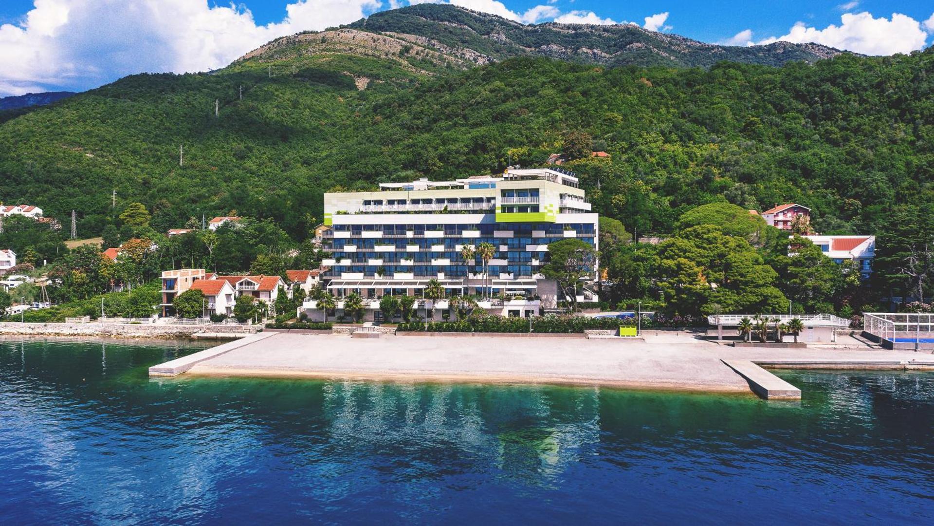Carine Hotel Park skats no jūras puses — viesnīcas ēka uz zaļu kalnu fona pie dzidra, zila ūdens Bijelā