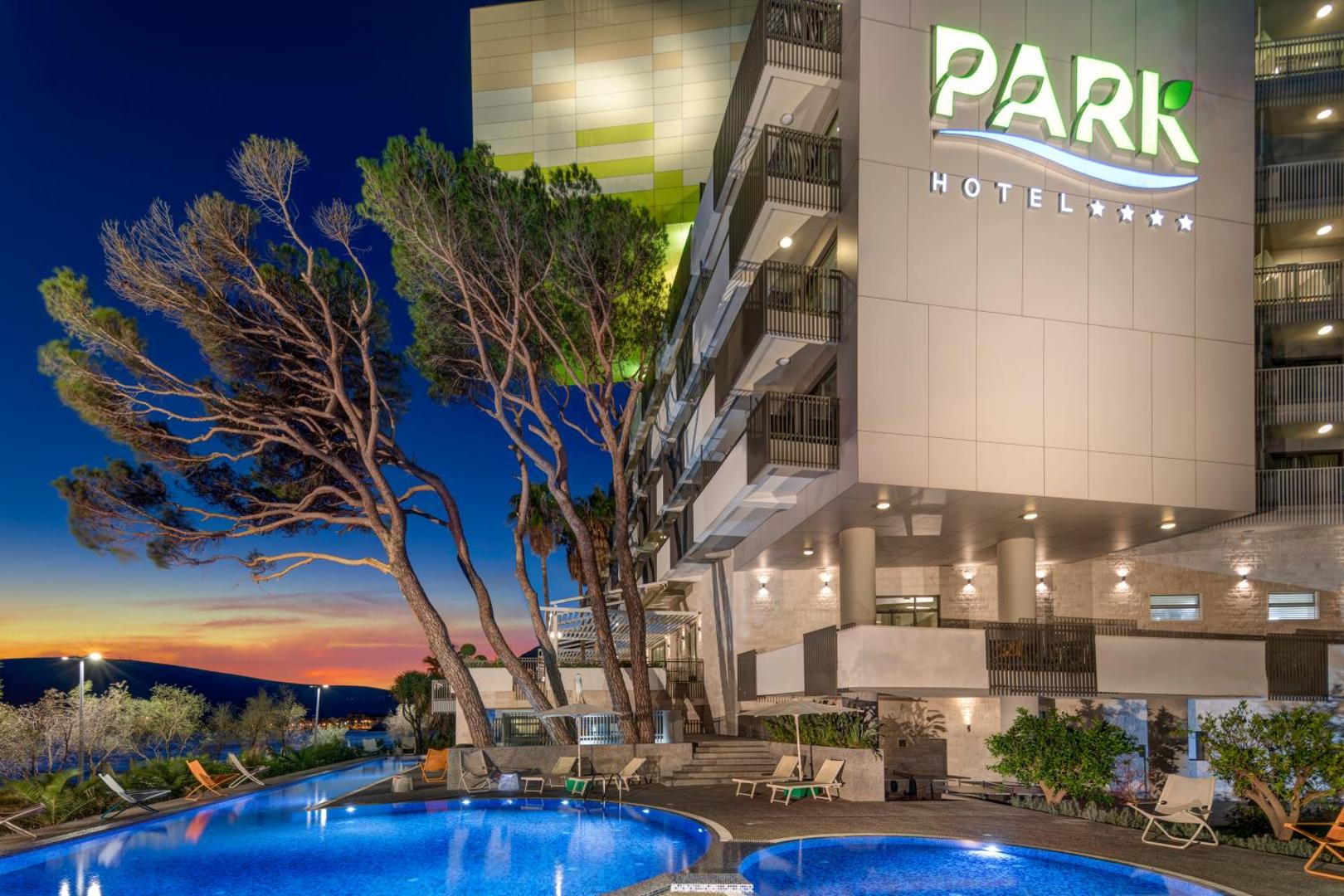 Carine Hotel Park fasāde un izgaismots āra baseins vakarā Bijelā, Melnkalnē — moderna arhitektūra ar balkoniem un priedēm saulrieta laikā