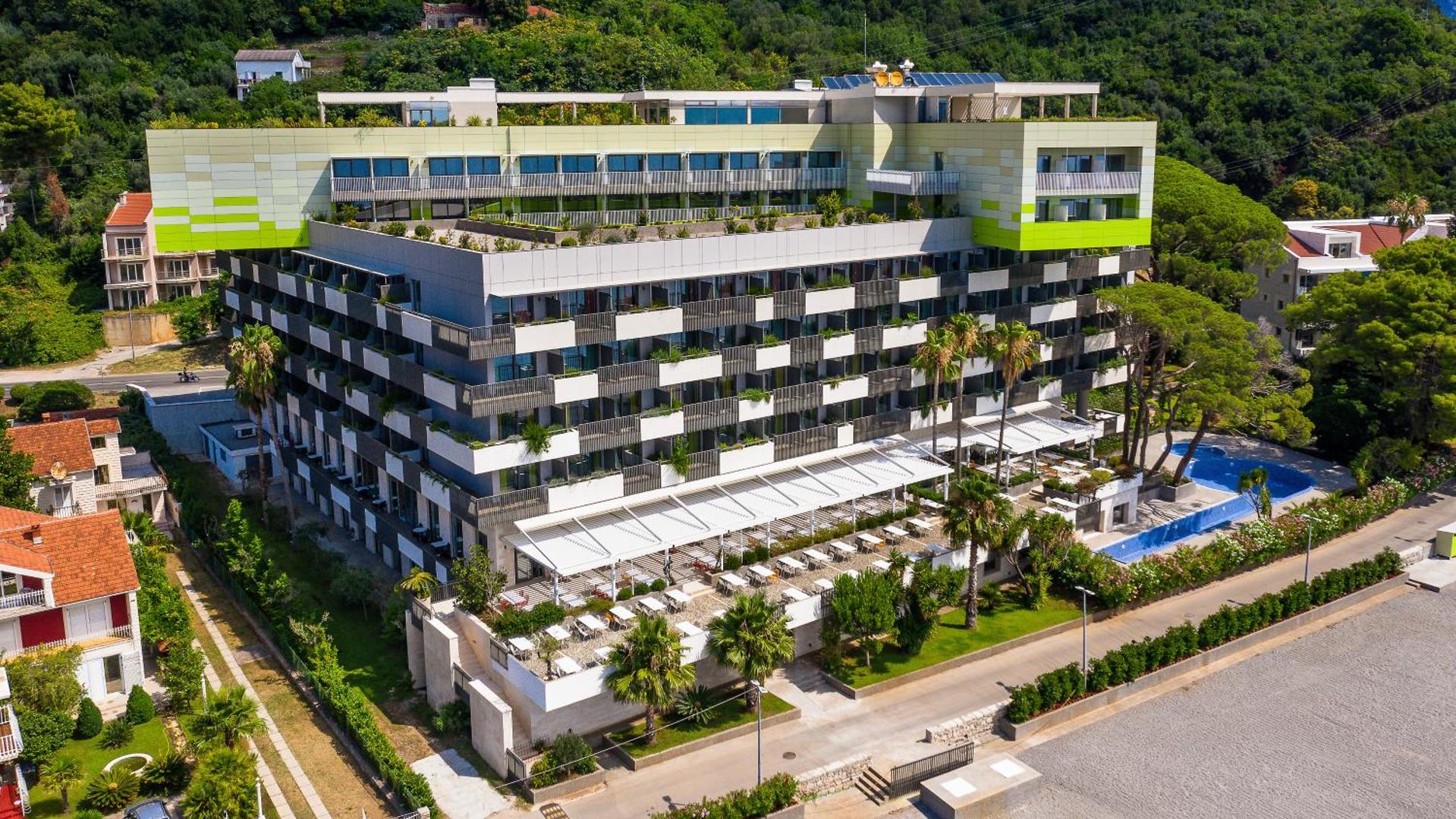 Carine Hotel Park modernā fasāde ar balkoniem, āra terasi un baseinu zonu, ko ieskauj zaļi koki Melnkalnes piekrastē