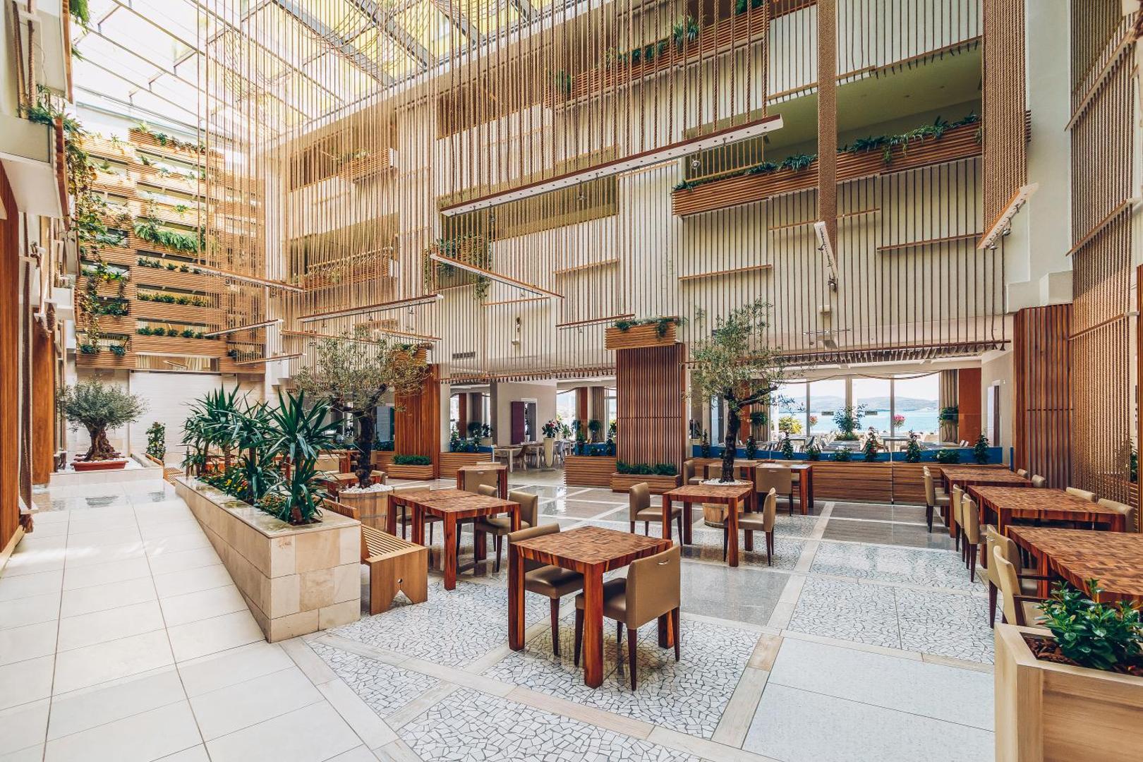 Carine Hotel Park modernais restorāns ar augstiem griestiem, koka apdari, dekoratīvām virvēm un dzīvajiem augiem iekštelpās