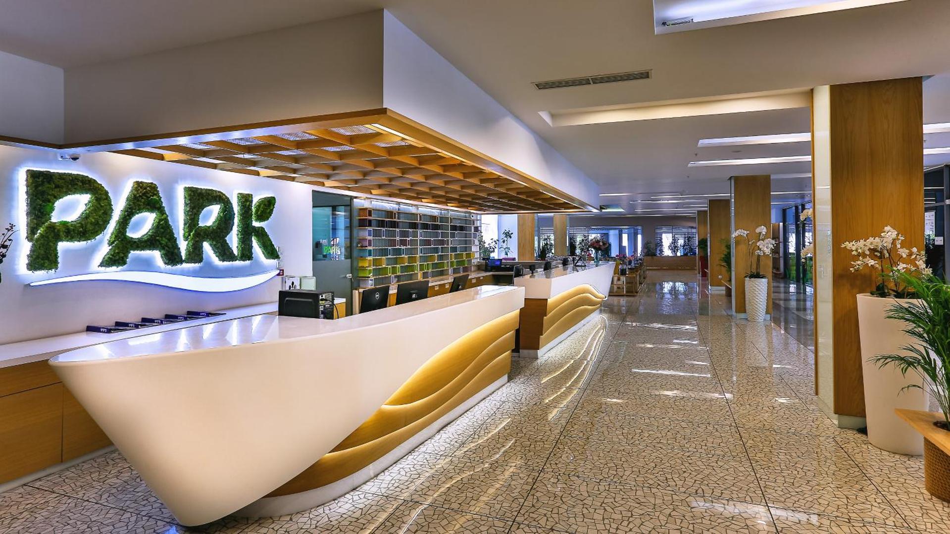 Carine Hotel Park modernā recepcijas zona ar gaišu, viļņveida dizaina leti un zaļojošu viesnīcas logo pie sienas