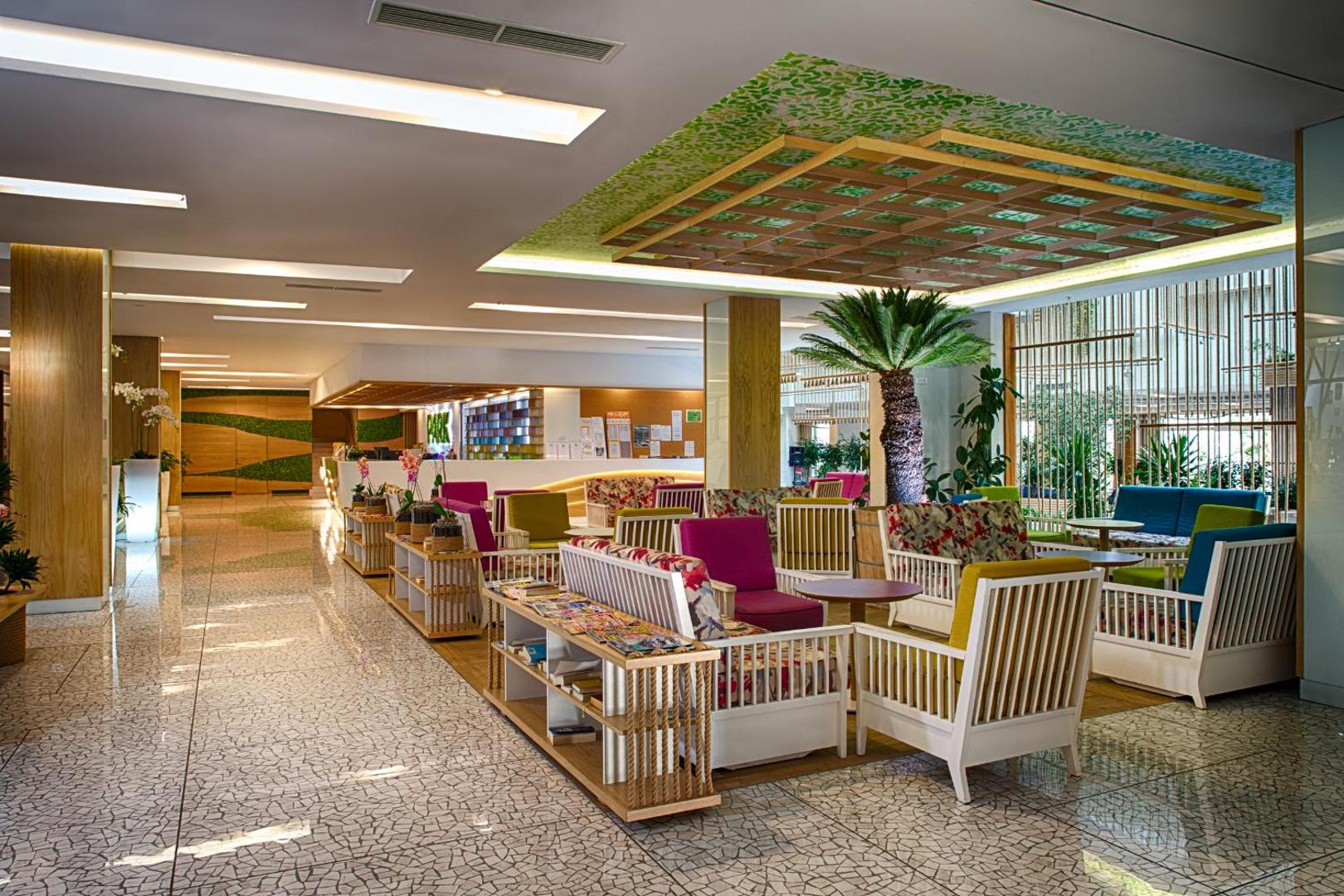 Carine Hotel Park lobby atpūtas zona ar krāsainiem atpūtas krēsliem, koka elementiem un dekoratīviem augiem gaišā interjerā