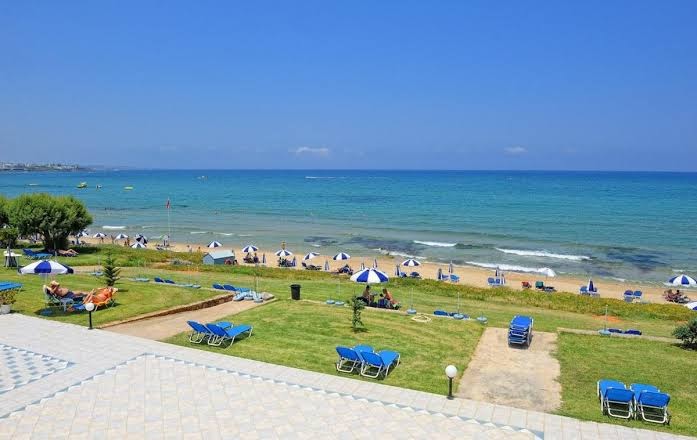 Ariadne Beach viesnīcas zaļā zona ar zālāju, sauļošanās krēsliem un skatu uz smilšu pludmali un Vidusjūru