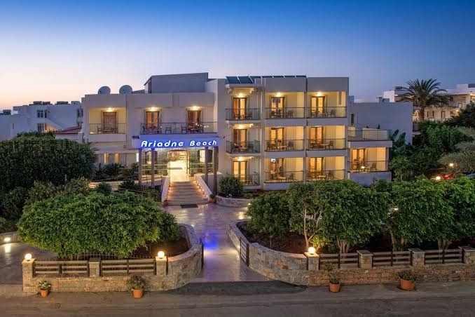 Ariadne Beach viesnīcas fasāde krēslas stundā ar izgaismotiem balkoniem, koptu dārzu un akmens mūra elementiem