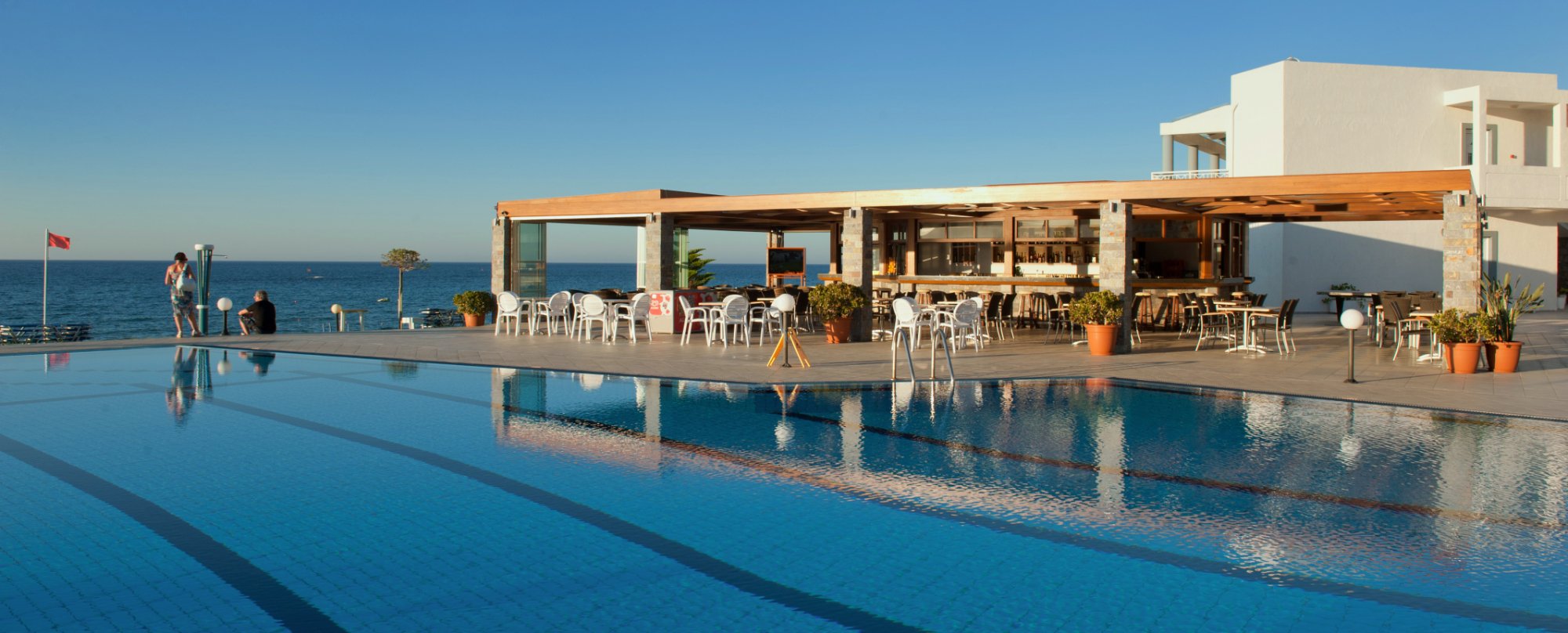 Ariadne Beach āra restorāns un bārs pie baseina ar skatu uz jūru saulrietā, balti galdi un krēsli uz terases