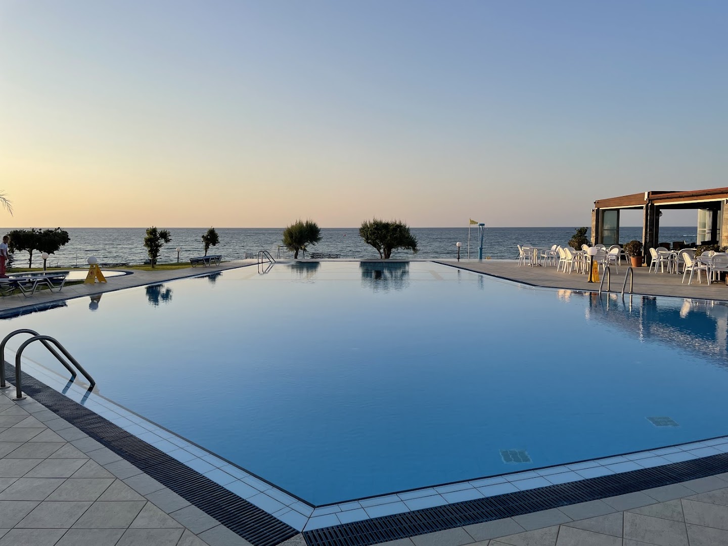 Liels, mierīgs Ariadne Beach baseins vakarā, kura ūdenī atspoguļojas debesis, blakus esošā terase un jūra tālumā