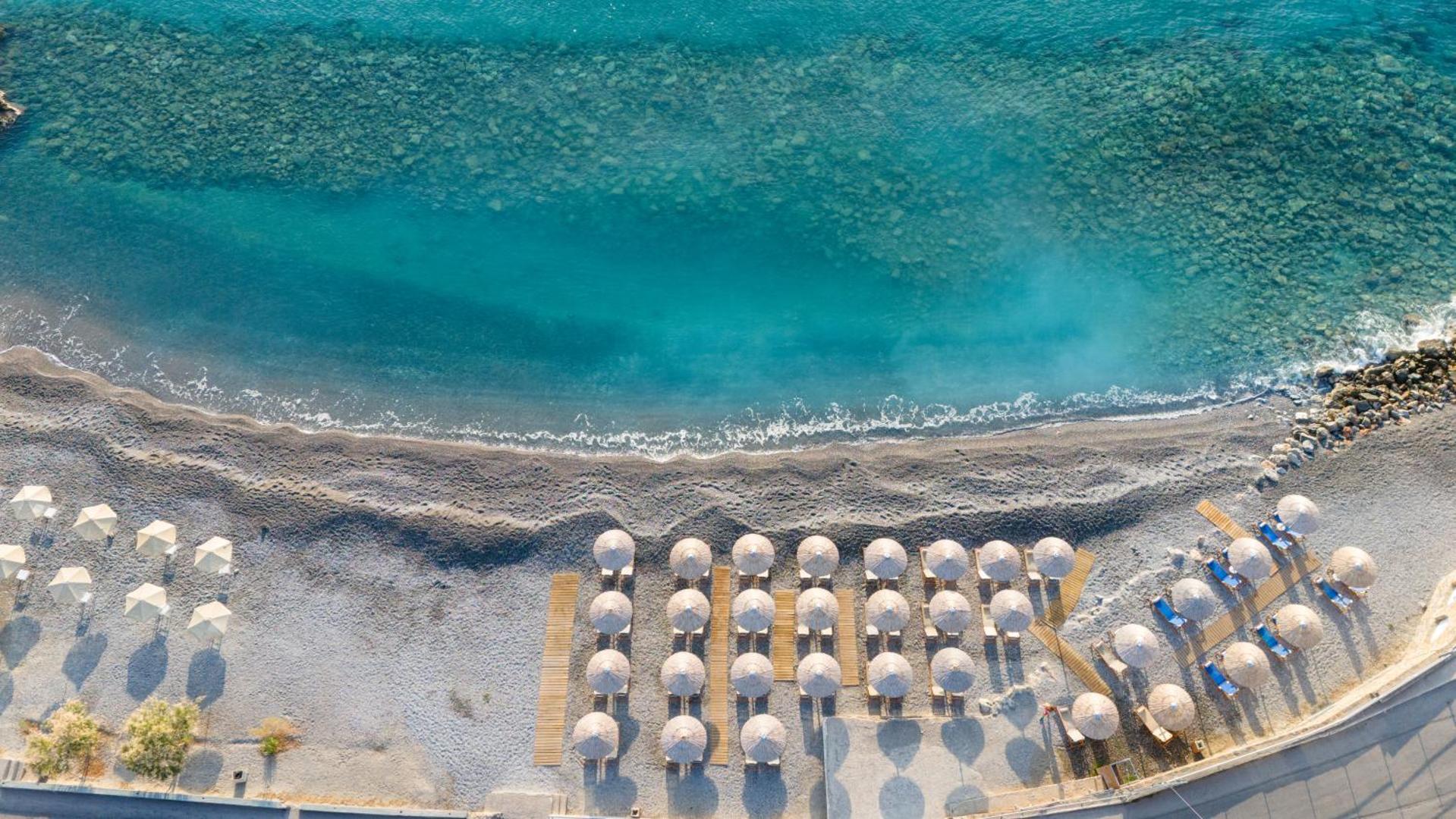 Ariadne Beach viesnīcas oļu pludmale no putna lidojuma Malijā, Krētā — tirkīzzils ūdens, saulessargi un sakopta krasta līnija