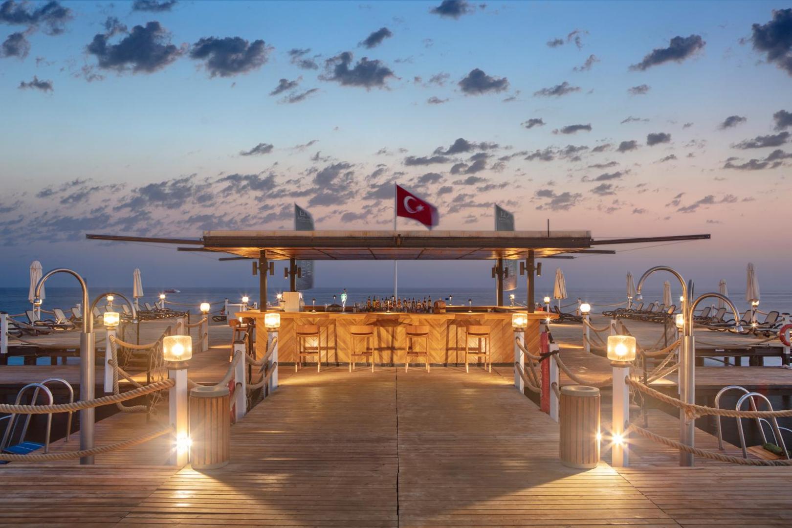 Ela Excellence Resort Belek bārs uz koka piestātnes jūrā saulrieta laikā — apgaismota lete ar krēsliem un Turcijas karogu