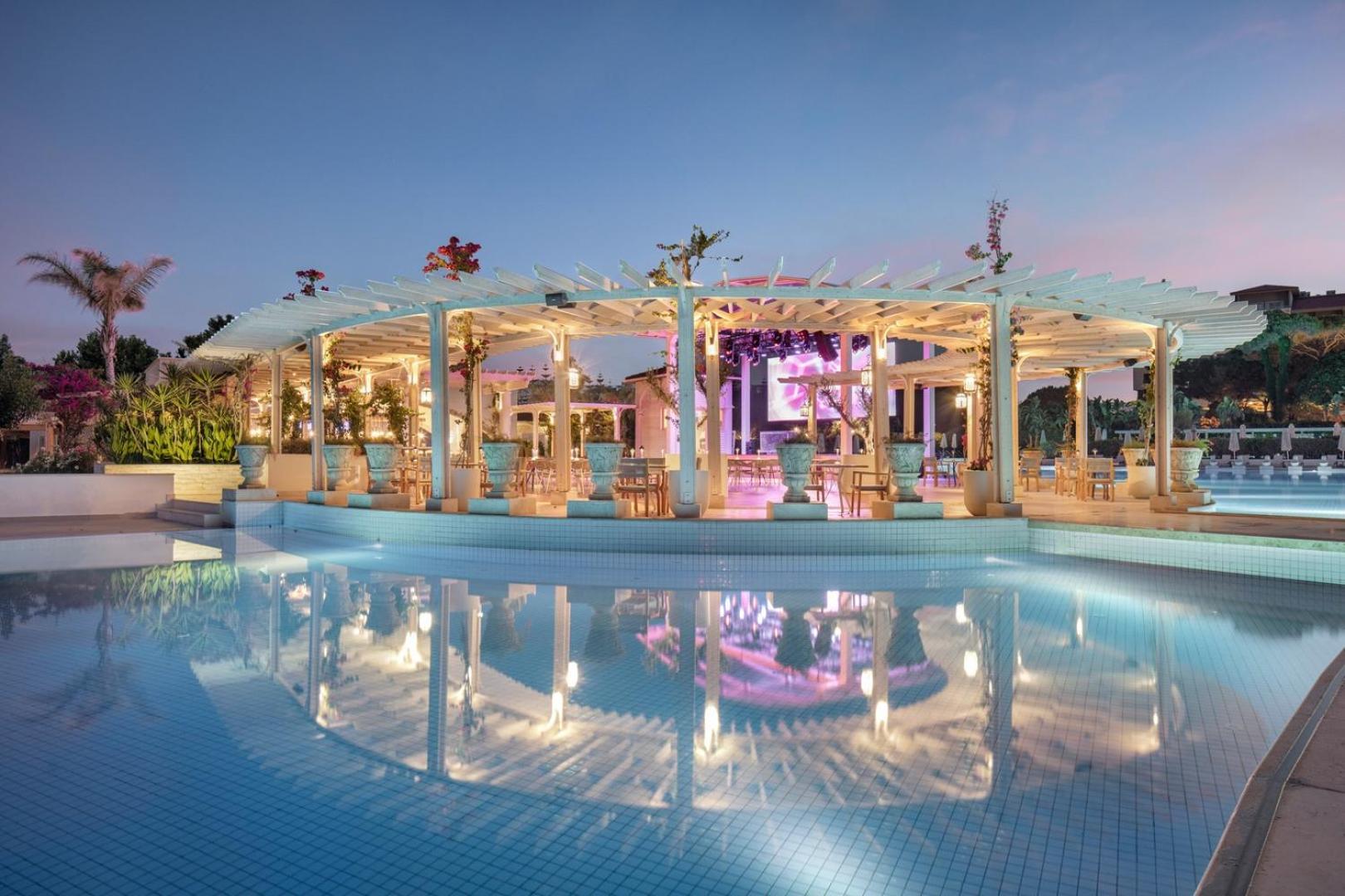 Ela Excellence Resort Belek āra baseins vakarā ar apgaismotu pergolu, restorāna zonu un atspulgu ūdenī Belekā.