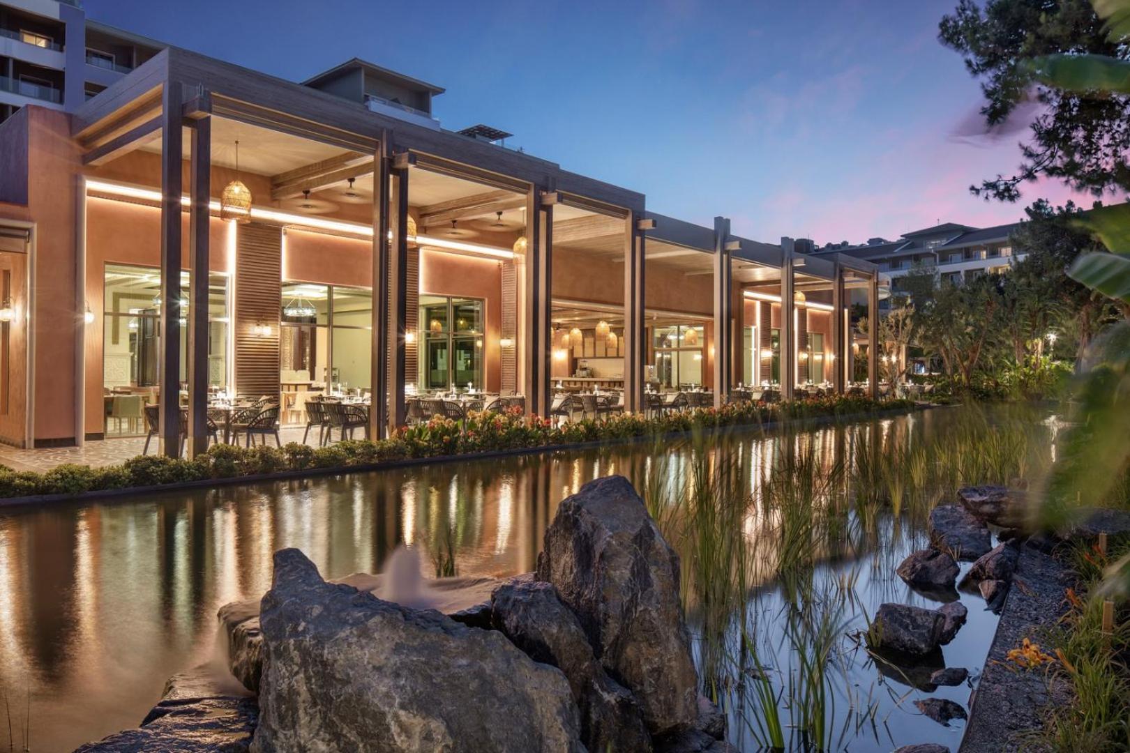 Ela Excellence Resort Belek modernā restorāna ēka pie dekoratīva ūdens kanāla saulrieta laikā Belekā, Turcijā