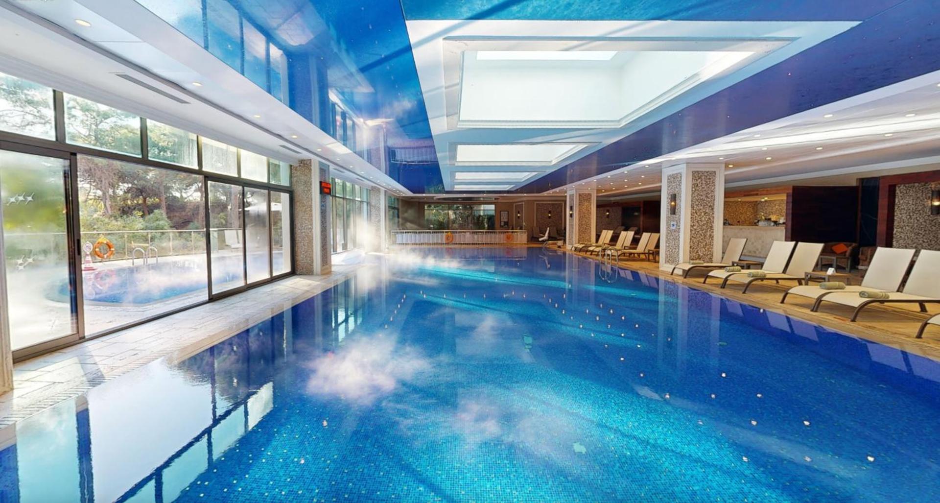 Ela Excellence Resort Belek iekštelpu baseins ar apsildi, panorāmas logiem un atpūtas krēsliem spa zonā Belekā, Turcijā