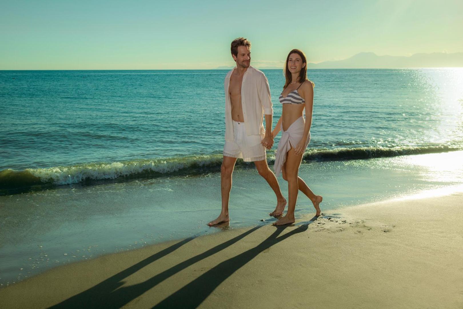 Ela Excellence Resort Belek privātā smilšu pludmale Turcijā — pāris pastaigājas gar jūras krastu saulrieta laikā zeltītos saules staros