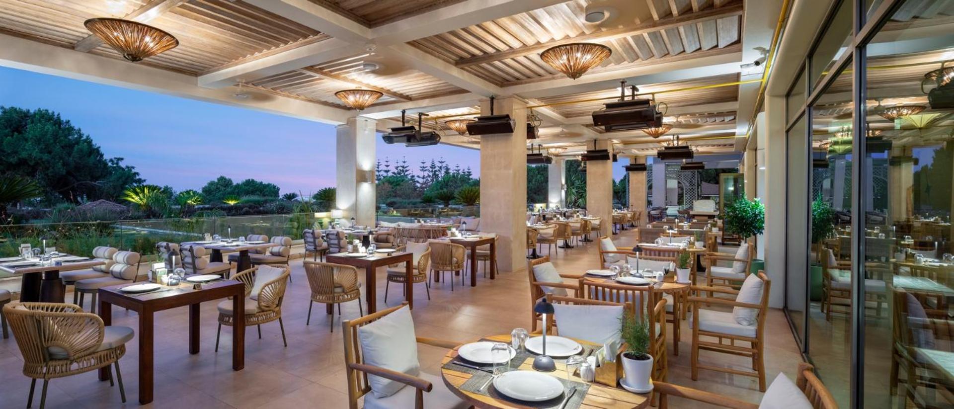 Ela Excellence Resort Belek restorāna terase vakarā ar elegantiem galdiņiem, pītiem krēsliem un siltu apgaismojumu Antaljas reģionā, Turcijā