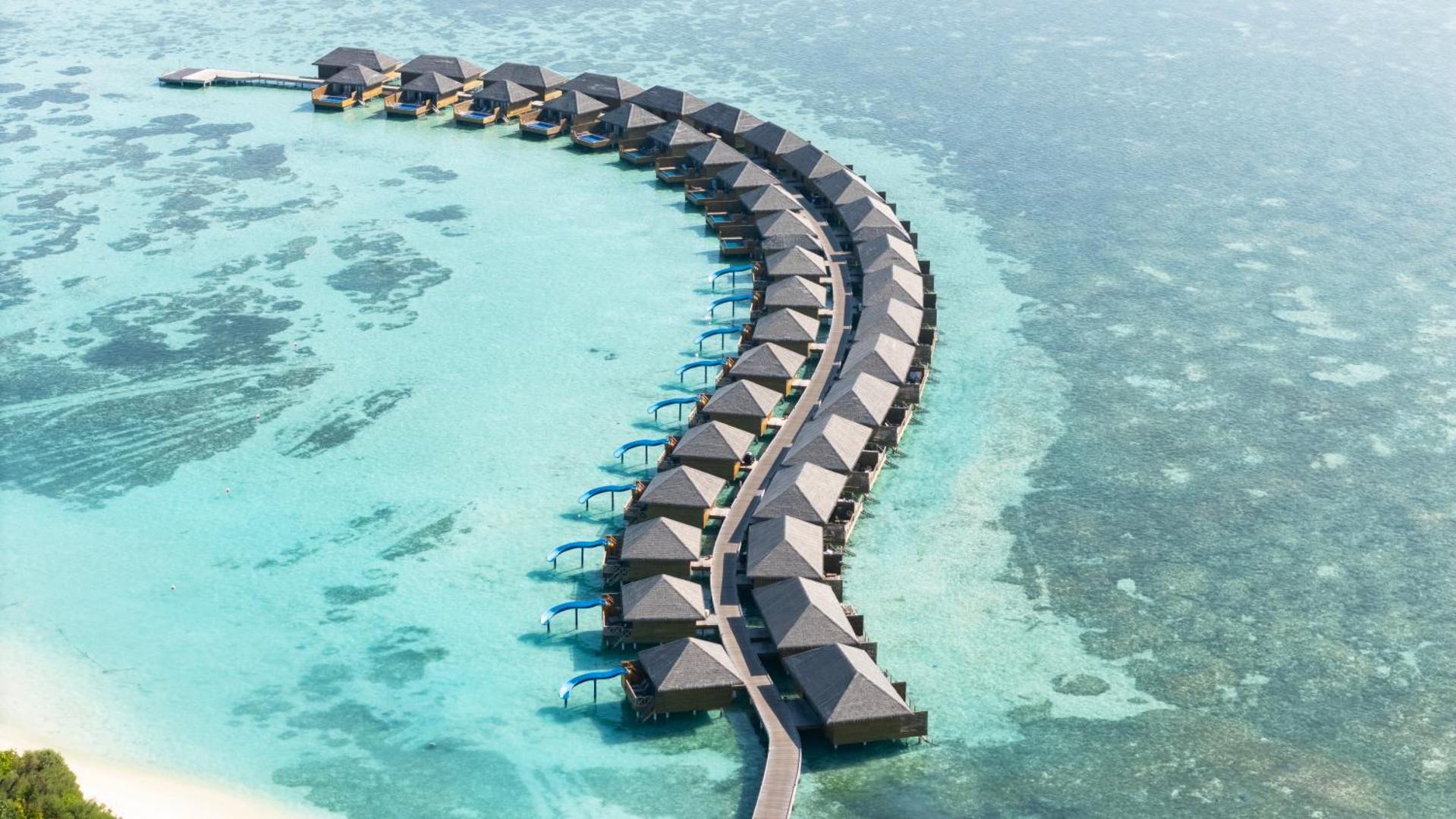 Cocoon Maldives ūdens bungalo rinda, kas izkārtota puslokā virs koraļļu rifa dzidrā Indijas okeāna ūdenī