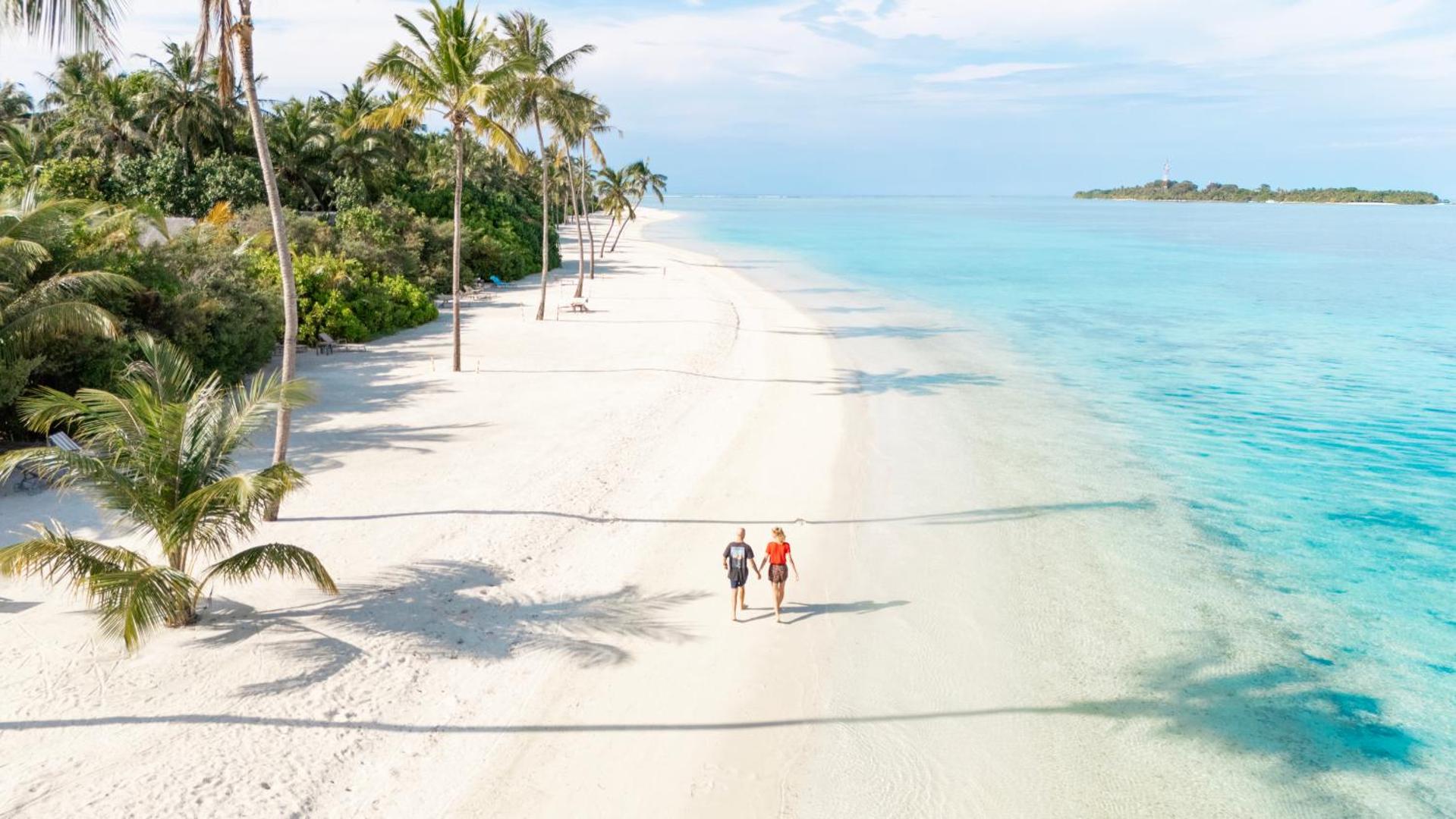Cocoon Maldives kūrorta sniegbaltā smilšu pludmale ar palmām un tirkīzzilu okeāna ūdeni — pāris pastaigājas gar krasta līniju Maldīvu salās