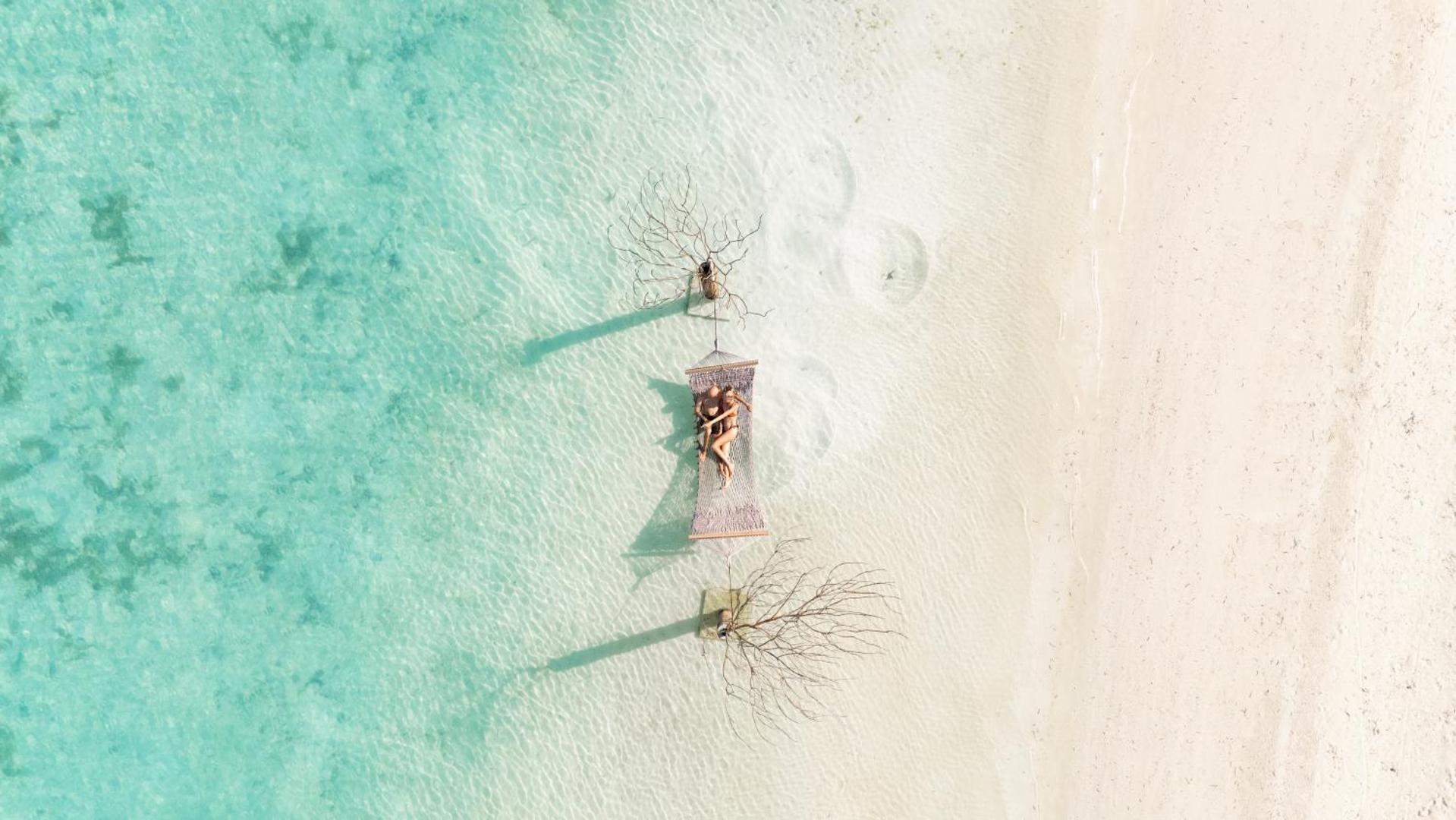 Skats no augšas uz Cocoon Maldives pludmali, kur starp diviem kokiem ūdens malā iekārts šūpuļtīkls ar atpūtniekiem