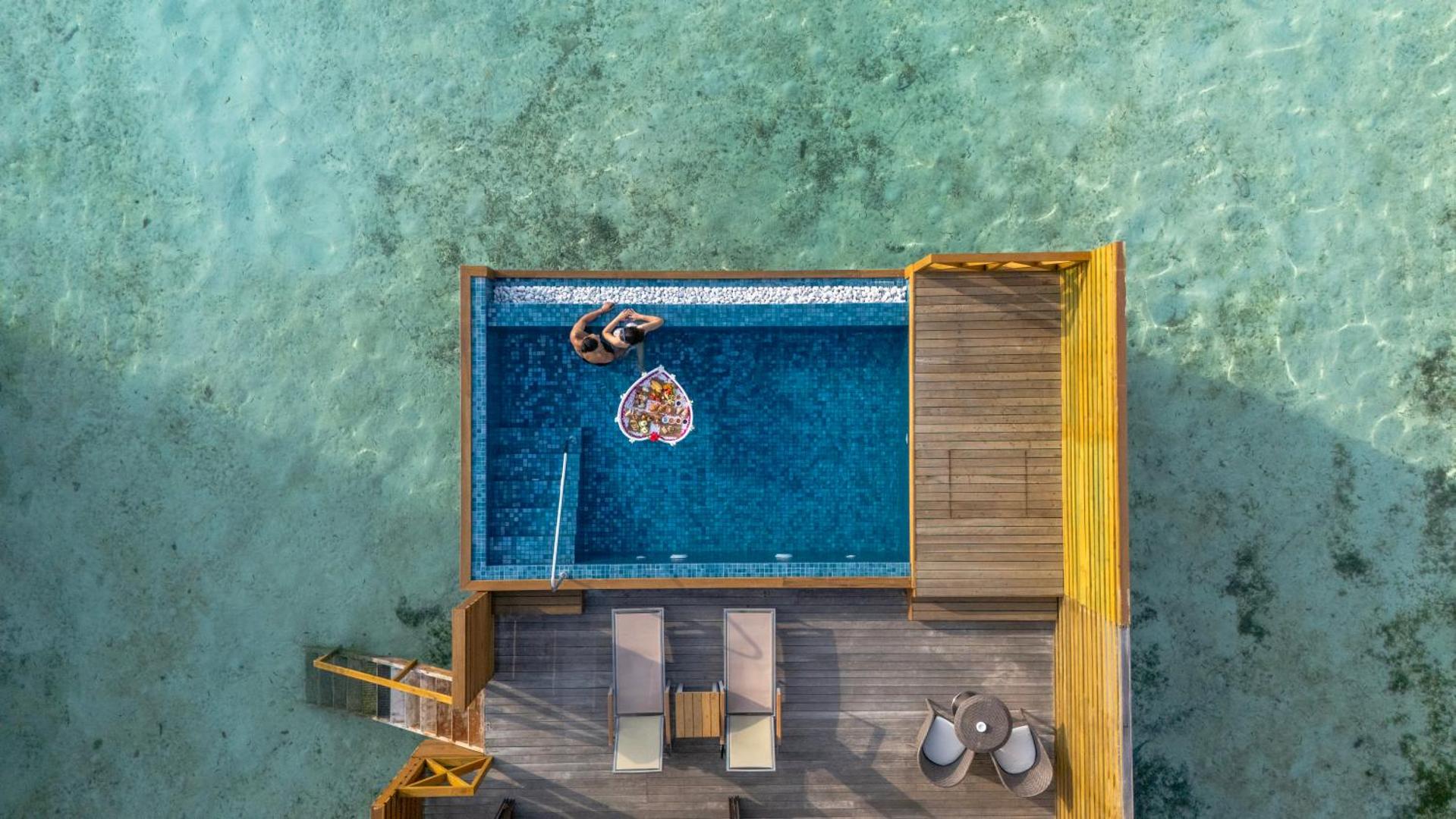 Cocoon Maldives kūrorta skats no augšas uz privāto baseinu virs ūdens ar peldošām brokastīm un pāri tirkīza lagūnā