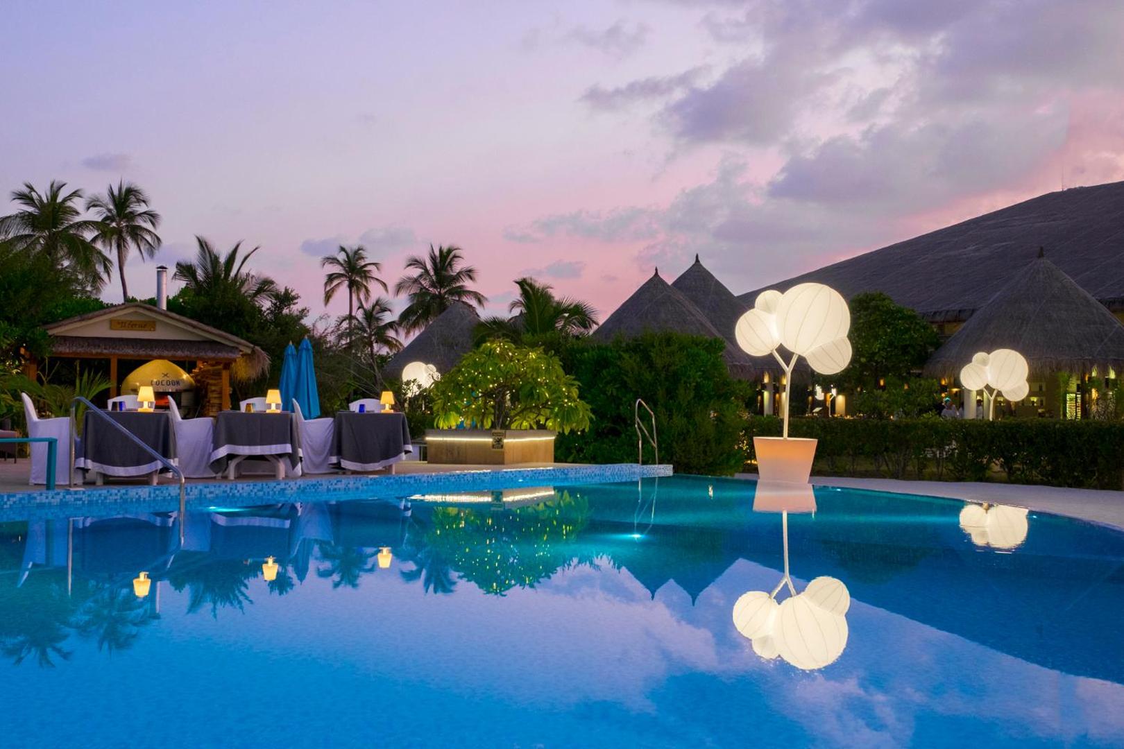 Cocoon Maldives vakara skats uz izgaismotu baseinu un restorāna terasi saulrieta laikā ar dekoratīvām gaismas instalācijām