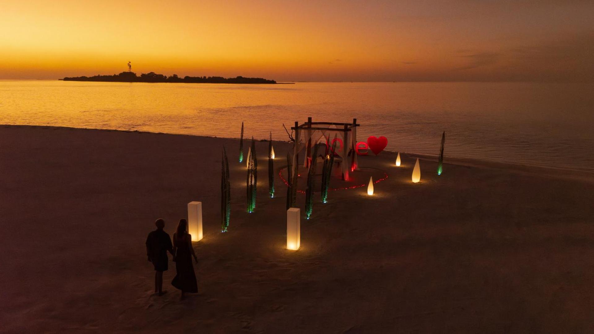 Romantiskas vakariņas Cocoon Maldives pludmalē saulrieta laikā — dekorēta nojume ar gaismām un sirds simbolu uz smiltīm pie okeāna