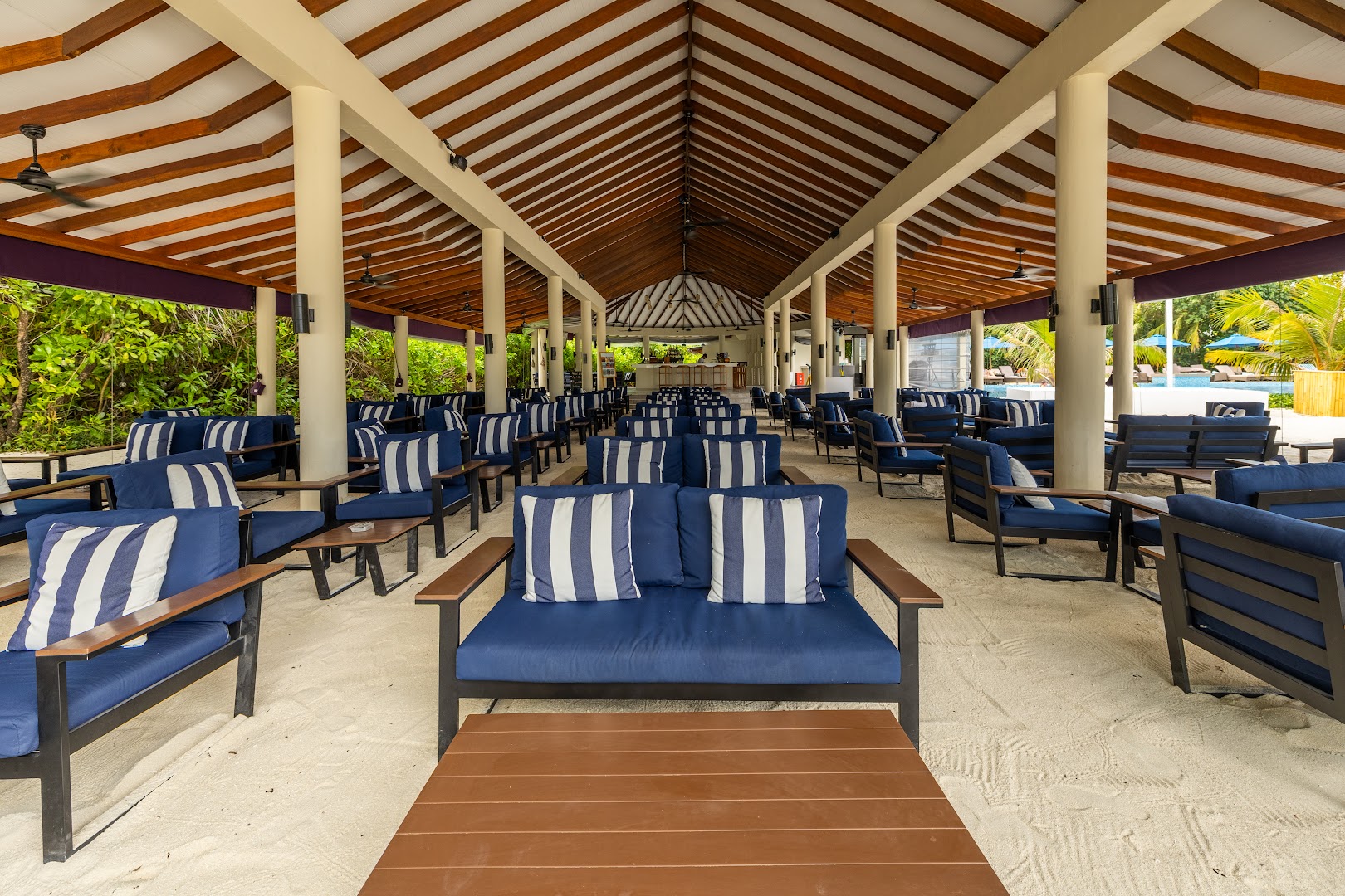 Cocoon Maldives plašā atpūtas zona ar ziliem dīvāniem un svītrainiem spilveniem uz smilšu grīdas zem koka jumta.