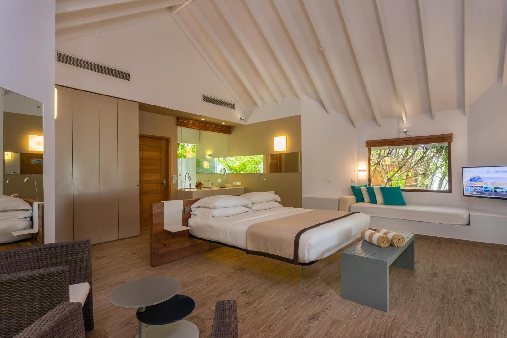 Cocoon Maldives moderns viesnīcas numurs ar gaišu koka apdari, plašu gultu un skatu uz dārzu caur logu