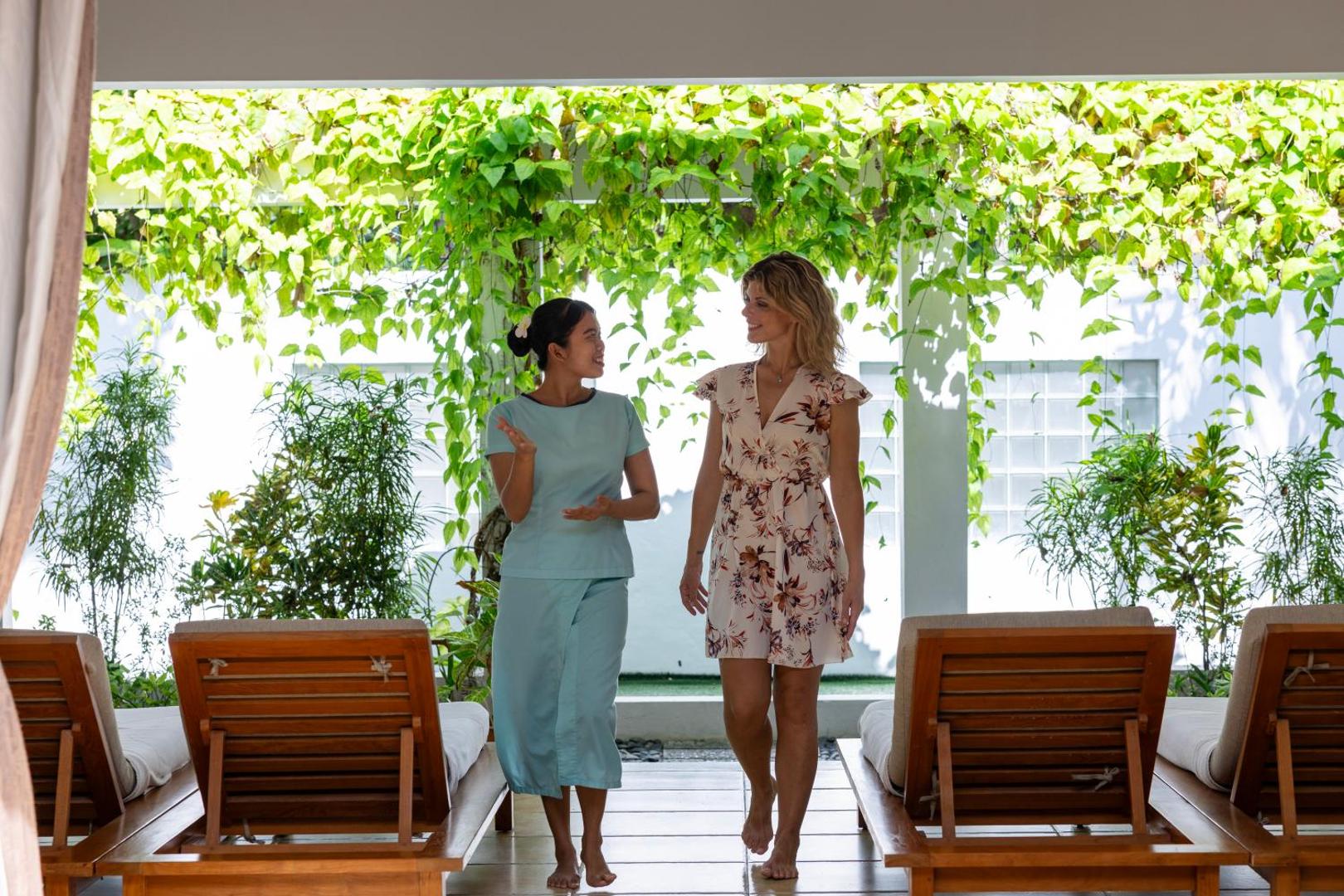 Viesnīcas darbiniece pavada viesi Cocoon Maldives spa zonā — koka sauļošanās krēsli un zaļojošu augu siena fonā