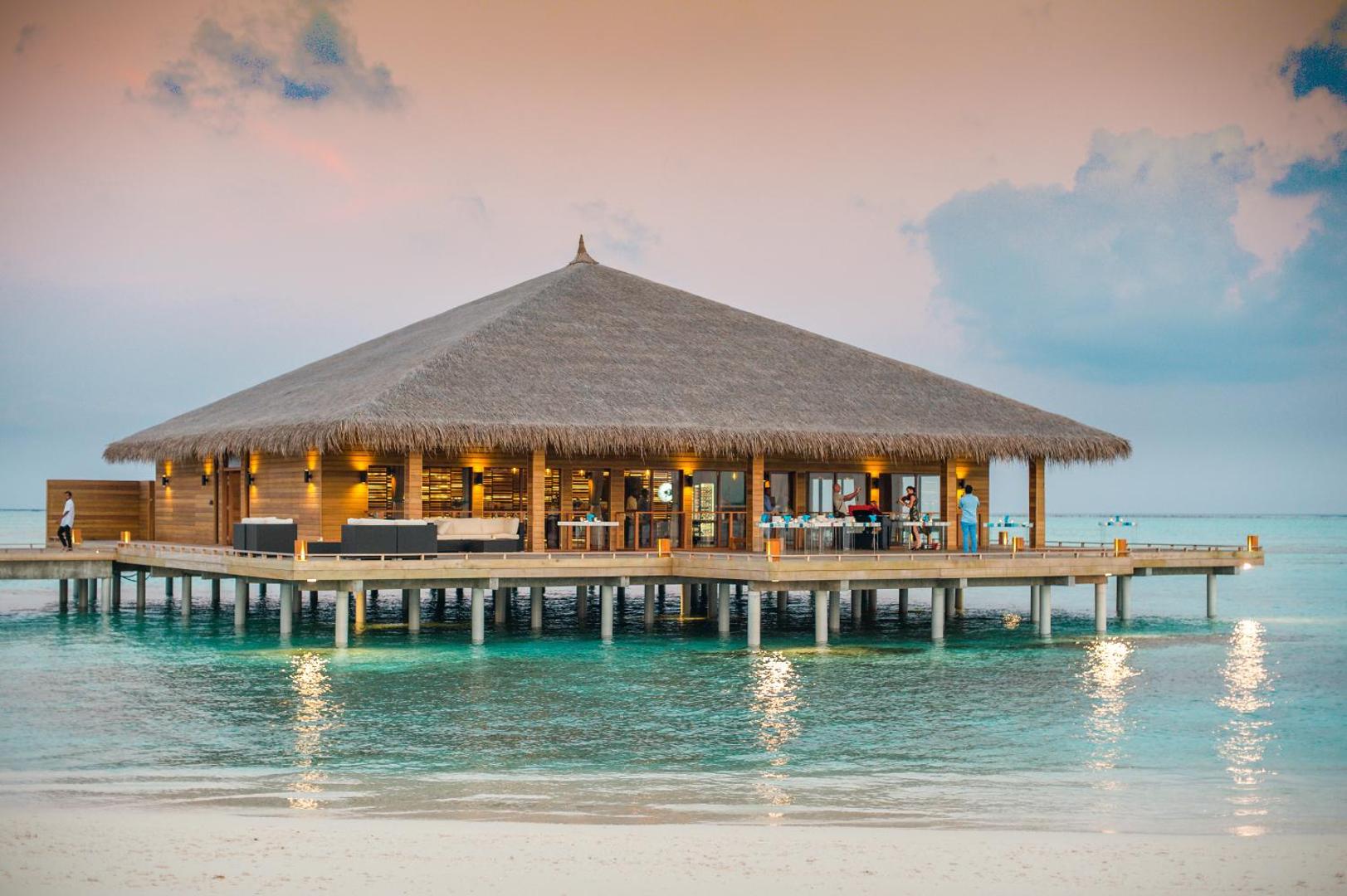 Cocoon Maldives restorāns uz pāļiem okeāna piekrastē saulrieta laikā — koka arhitektūra ar salmu jumtu un apgaismotu terasi