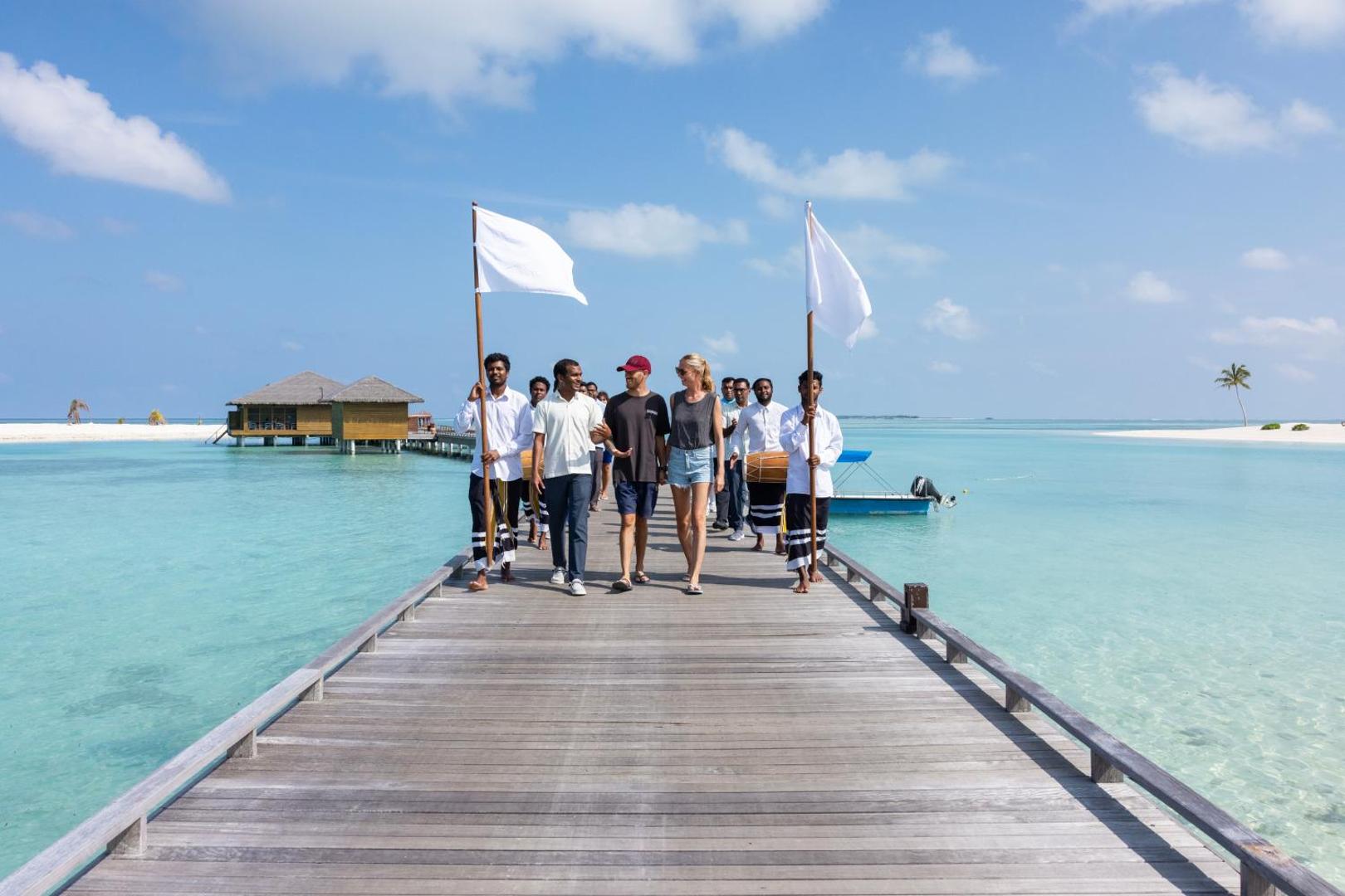 Viesu sagaidīšanas ceremonija uz Cocoon Maldives koka laipas virs tirkīzzila ūdens ar tradicionālo mūziku un karogiem.