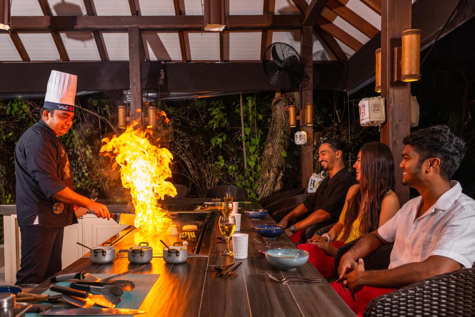 Cocoon Maldives šefpavārs gatavo ēdienu uz atklātas uguns Teppanyaki restorānā, viesiem vērojot kulinārijas šovu
