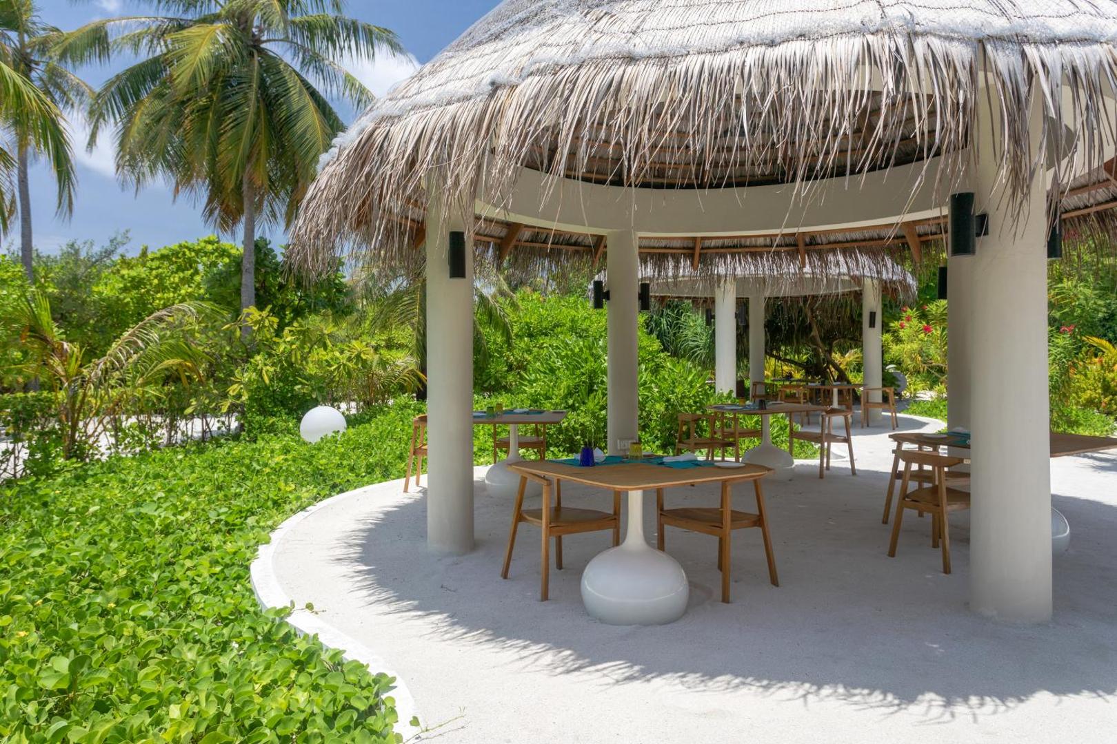 Cocoon Maldives āra restorāns zem salmu jumta nojumes dārza zonā ar koka galdiem un krēsliem uz baltām smiltīm