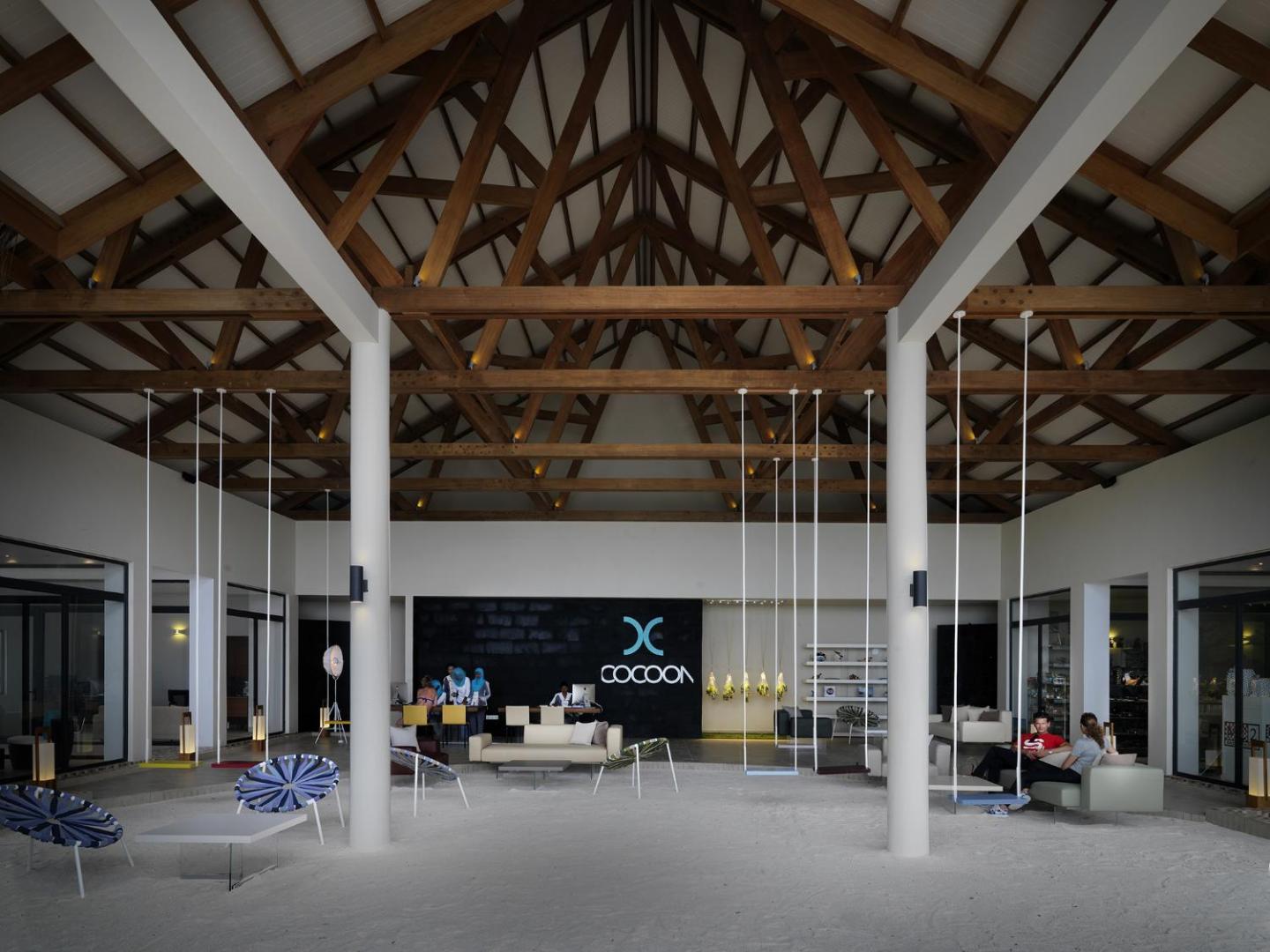 Cocoon Maldives viesnīcas lobby ar augstiem koka griestiem un smilšu grīdu — atpūtas zona ar šūpolēm un modernām mēbelēm