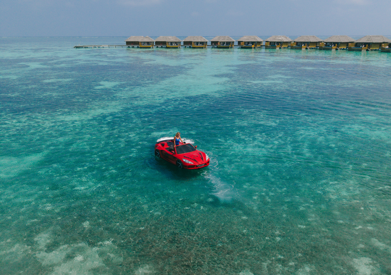 Aerofoto ar Cocoon Maldives ūdens bungalo un sarkanu ūdens transportlīdzekli dzidrā, tirkīzzilā Indijas okeāna ūdenī.
