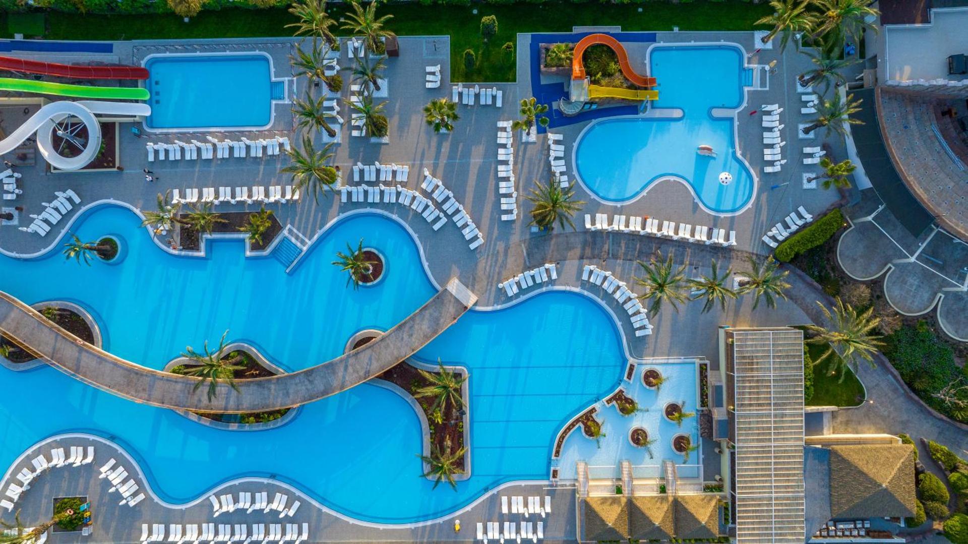 Liberty Hotels Lara baseinu komplekss no putna lidojuma Antālijā — plaša atpūtas zona ar vairākiem baseiniem, tiltiņiem, palmām un ūdens slidkalniņiem