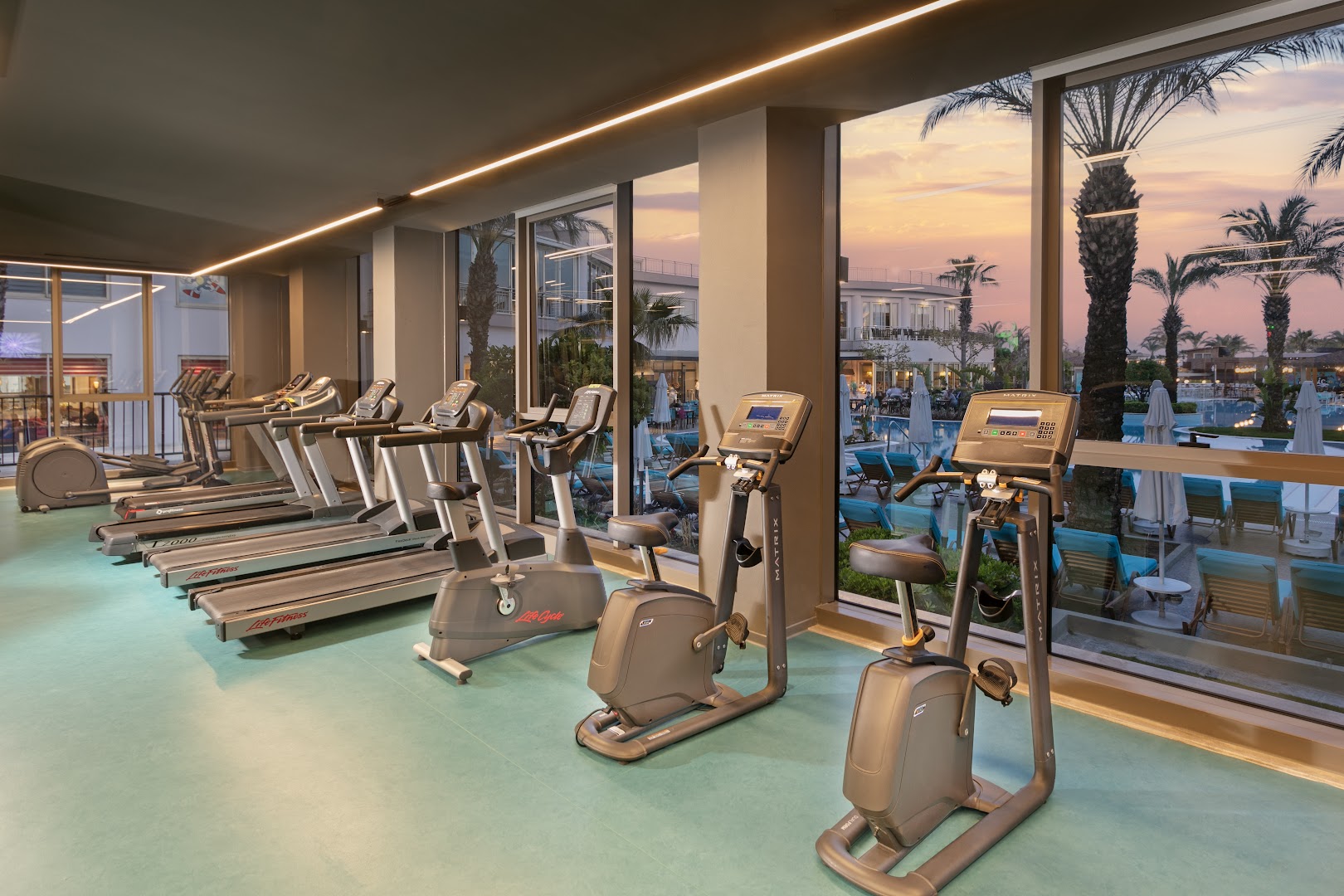 Liberty Hotels Lara modernais fitness centrs ar skrejceliņiem un velotrenažieriem — lieli logi ar skatu uz baseinu un palmām