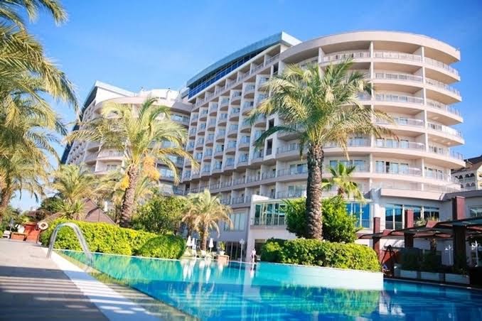 Skats uz Liberty Hotels Lara moderno arhitektūru no baseina malas, ko rotā augstas palmas un zaļojoši krūmi