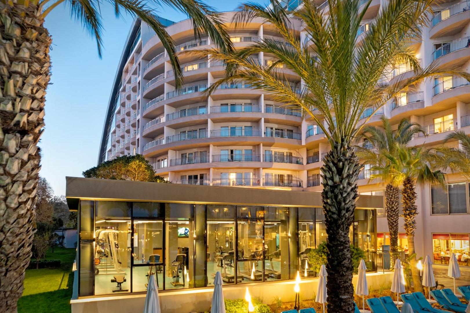Liberty Hotels Lara viesnīcas ēkas fasāde un fitness centrs vakarā — palmas, apgaismoti balkoni un stiklota trenažieru zāle